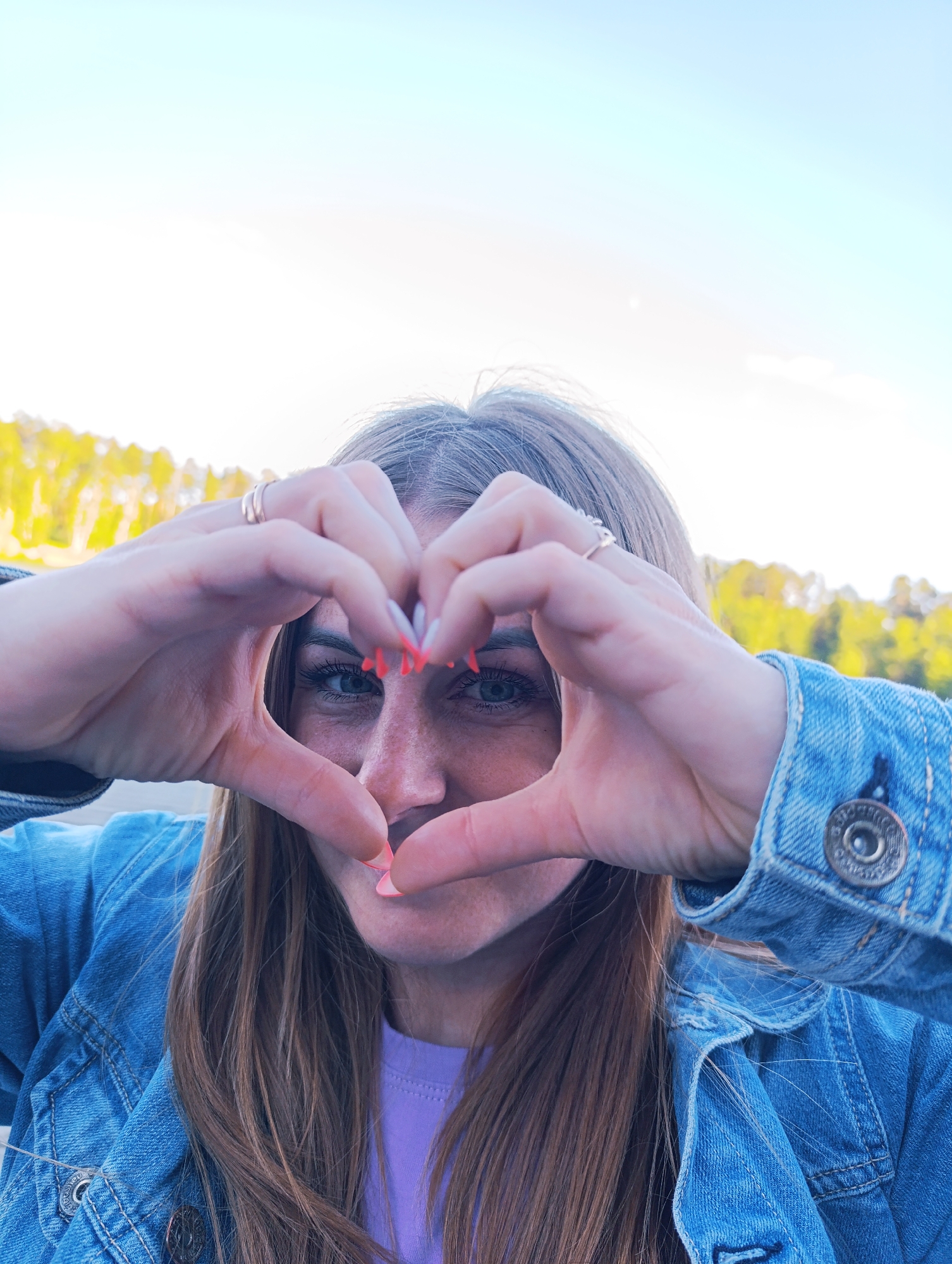 A young woman is pictured from the chest up, making a heart shape with her hands in front of her face. She has long, light brown hair that falls past her shoulders. She is wearing a blue denim jacket over a purple t-shirt. Her fingernails are painted a bright coral color. She has striking blue eyes and a smattering of freckles across her nose and cheeks. One ring is visible on her left hand, and another on her right index finger.
The background shows a blurred landscape of trees with yellow foliage, suggesting it is autumn. The sky above is a pale blue, indicating it is daytime, likely late afternoon due to the warm lighting. The overall mood of the image is positive and affectionate. There is no visible text in the image.