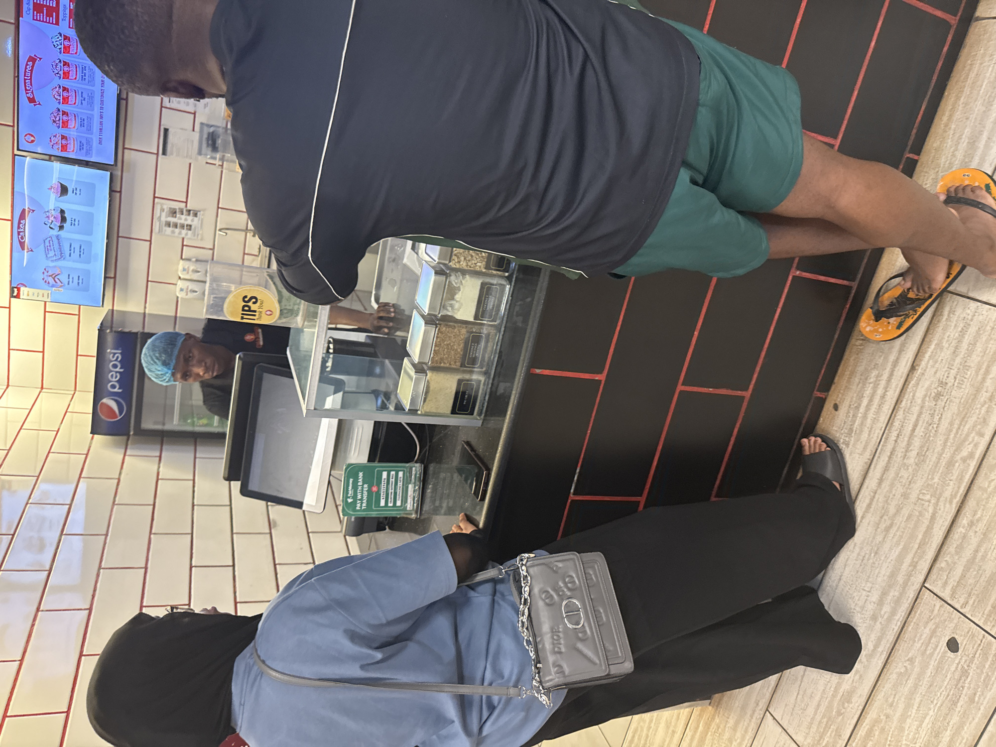 People stand at a food service counter. A man wearing a black shirt and green shorts waits to be served. He is wearing flip-flops. A woman in a blue shirt and a hijab stands behind him. She wears black pants and sandals and carries a gray purse. Behind the counter, a person with a blue hair covering fills the order. A sign says, "Tips, Thank You!". A Pepsi sign is visible. Digital menus are visible with descriptions of cakes, signatures, cap sizes, and toppings. A sign says, "Pay with bank transfer". The location is likely a sweets shop or cafe in Abuja, Nigeria. The walls are white and the floor is beige wood-look planks.