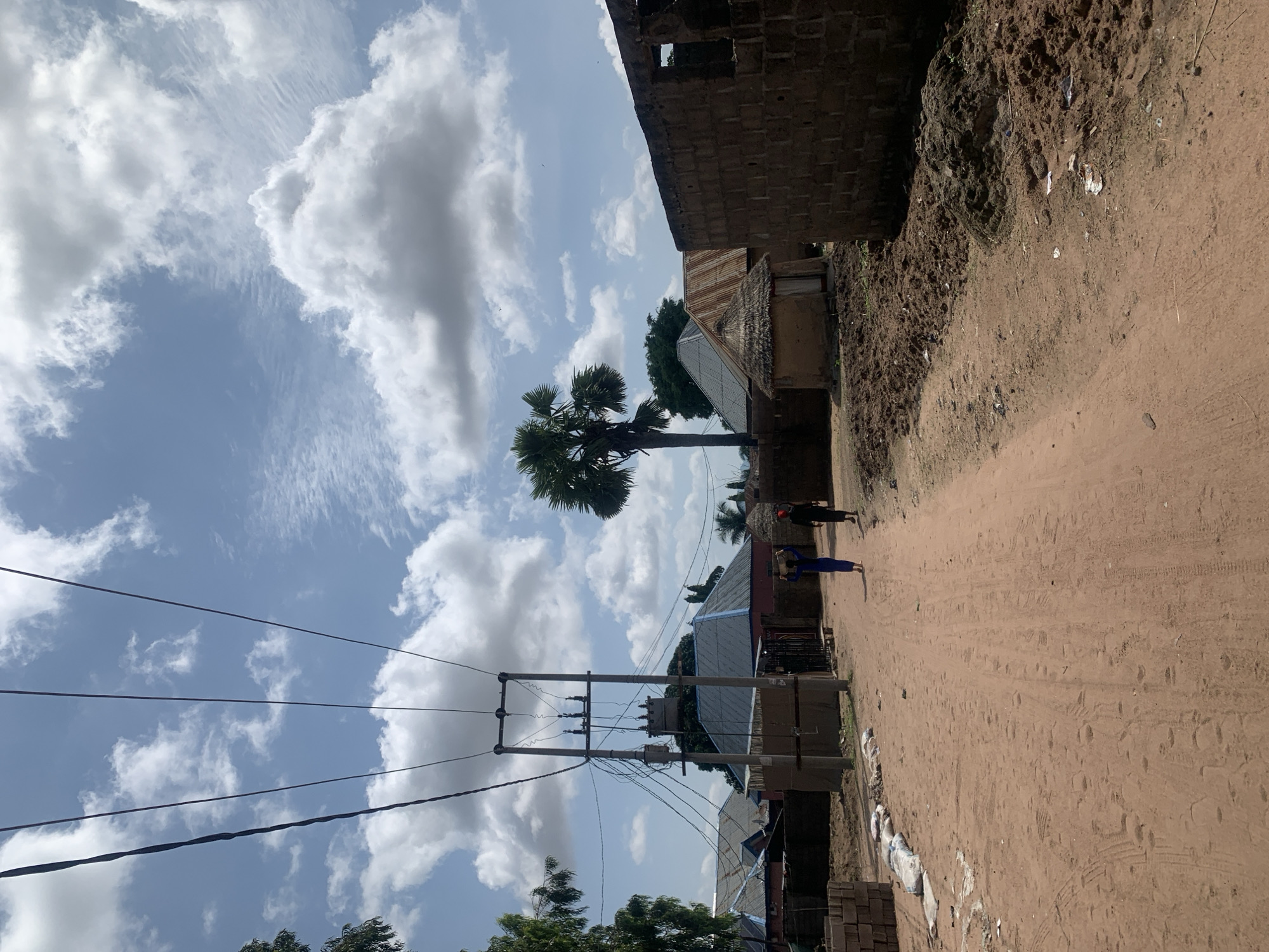 The image depicts a dusty dirt road in a village setting, likely in Jalingo, Nigeria. The road is flanked by various structures, including houses with corrugated metal roofs and some made of mud with thatched roofs. In the foreground, stacks of bricks and bags of what appear to be cement or sand are visible on the left side of the road. Further down the road, two individuals are walking away from the camera. The person in front is wearing a blue garment and carrying a basket on their head, while the person behind them is wearing dark clothing and a red head covering. A tall palm tree stands prominently in the background, with more trees and vegetation scattered throughout the scene. Overhead, a utility pole with numerous wires stretches across the sky, and a transformer is visible on the pole. The sky is partially cloudy, with fluffy white clouds against a blue backdrop. The overall impression is one of a rural community in a warm, sunny climate.