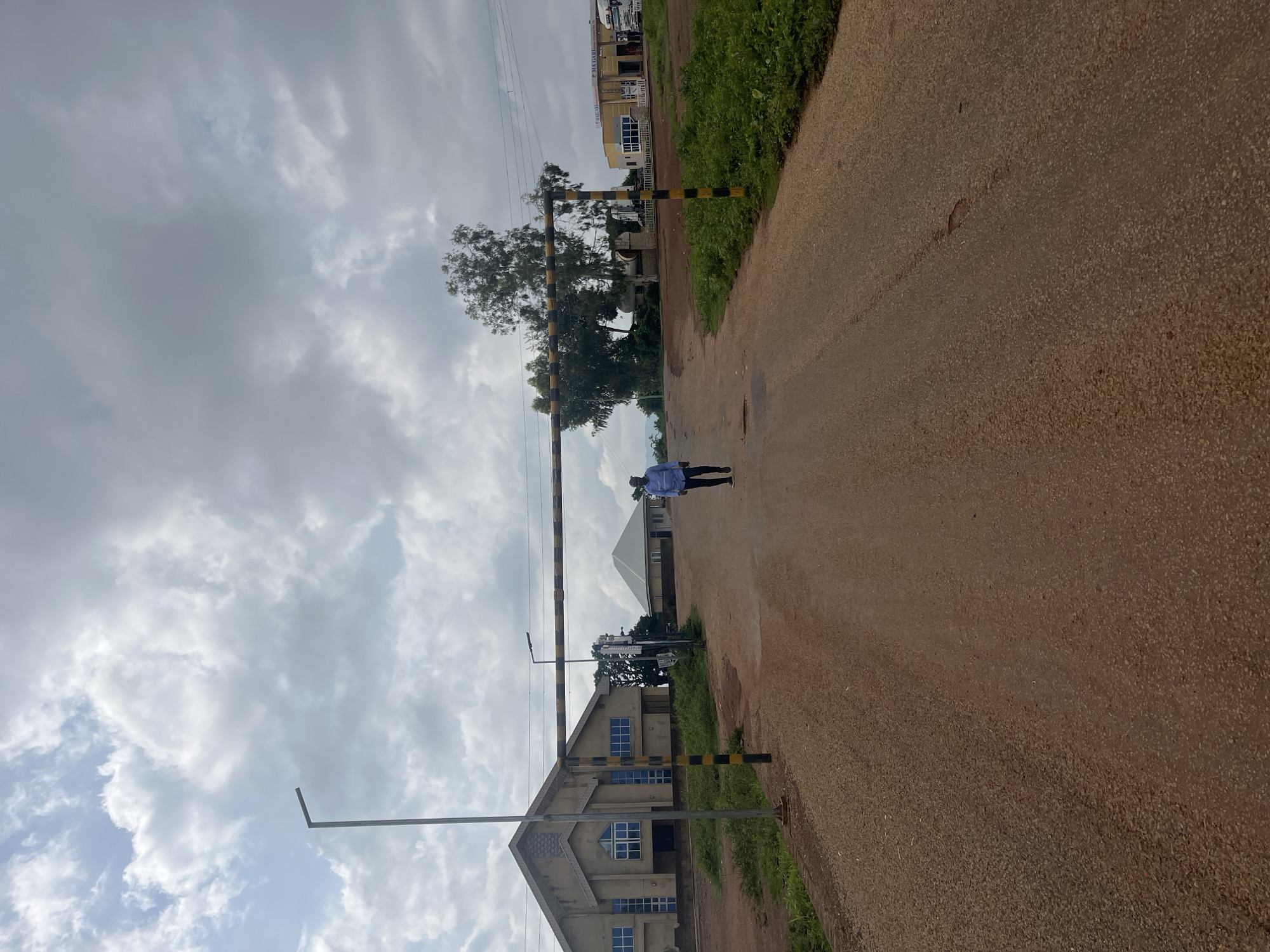 In Unguwar Saya, Nigeria, a person walks down a dirt road towards a building with a pitched roof. A large, beige building with blue-windowed facades is visible on the left, and another yellow building is to the right. A barrier with yellow and black stripes is suspended horizontally across the road, supported by posts. The sky is cloudy.