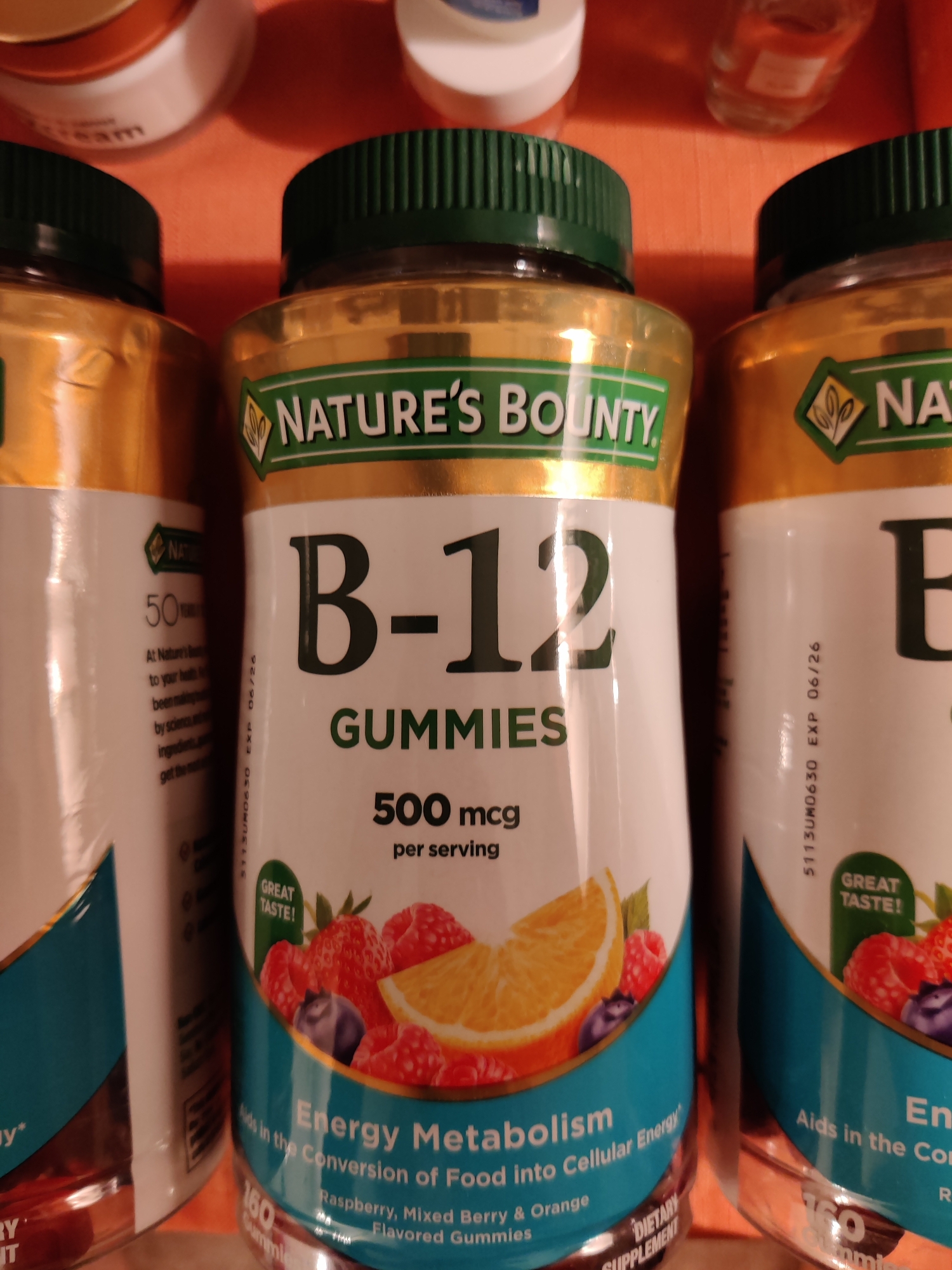 Three bottles of Nature's Bounty B-12 Gummies are arranged in the foreground of the image, suggesting a shelf or countertop in a retail or home environment. The bottles are mostly white with gold accents, featuring a green lid and a green label with white text and a green nature-themed logo. The central bottle prominently displays "B-12 GUMMIES" in large, dark gray lettering, followed by "500 mcg per serving" and an illustration of assorted berries and citrus fruit. A "GREAT TASTE!" banner is also visible on this bottle. The leftmost bottle has a partially visible label that includes "50 YEARS" and text describing the product. The rightmost bottle is similar to the central one, with "B-" clearly visible as the start of the product name. Behind the bottles, out of focus, are the tops of other containers, one with a gold lid and cream-colored base, and another white container. The background appears to be an orange surface. The lighting is somewhat dim, with a slight overhead source casting subtle shadows. No people or animals are present. The location in Lower Paxton Township, United States of America, is inferred from general knowledge about the product's availability rather than any visual cues in the image.