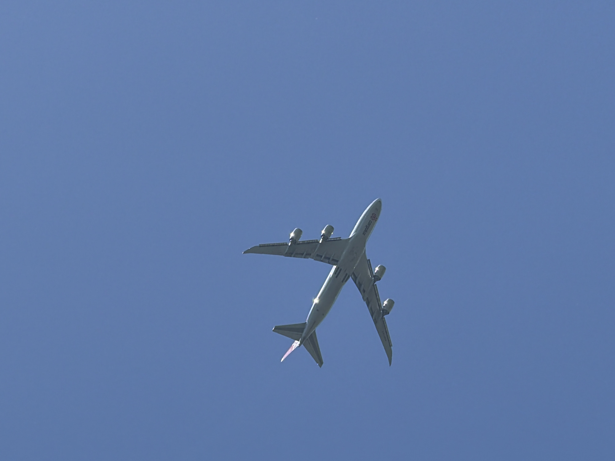 A Cargolux Boeing 747-400F airplane is flying in the clear blue sky above the city of Pétange, Luxembourg. The airplane is ascending, with its undercarriage not visible, its four engines apparent beneath each wing, and the Cargolux logo visible on its fuselage. The tail fin is directed towards the lower-left corner of the image.