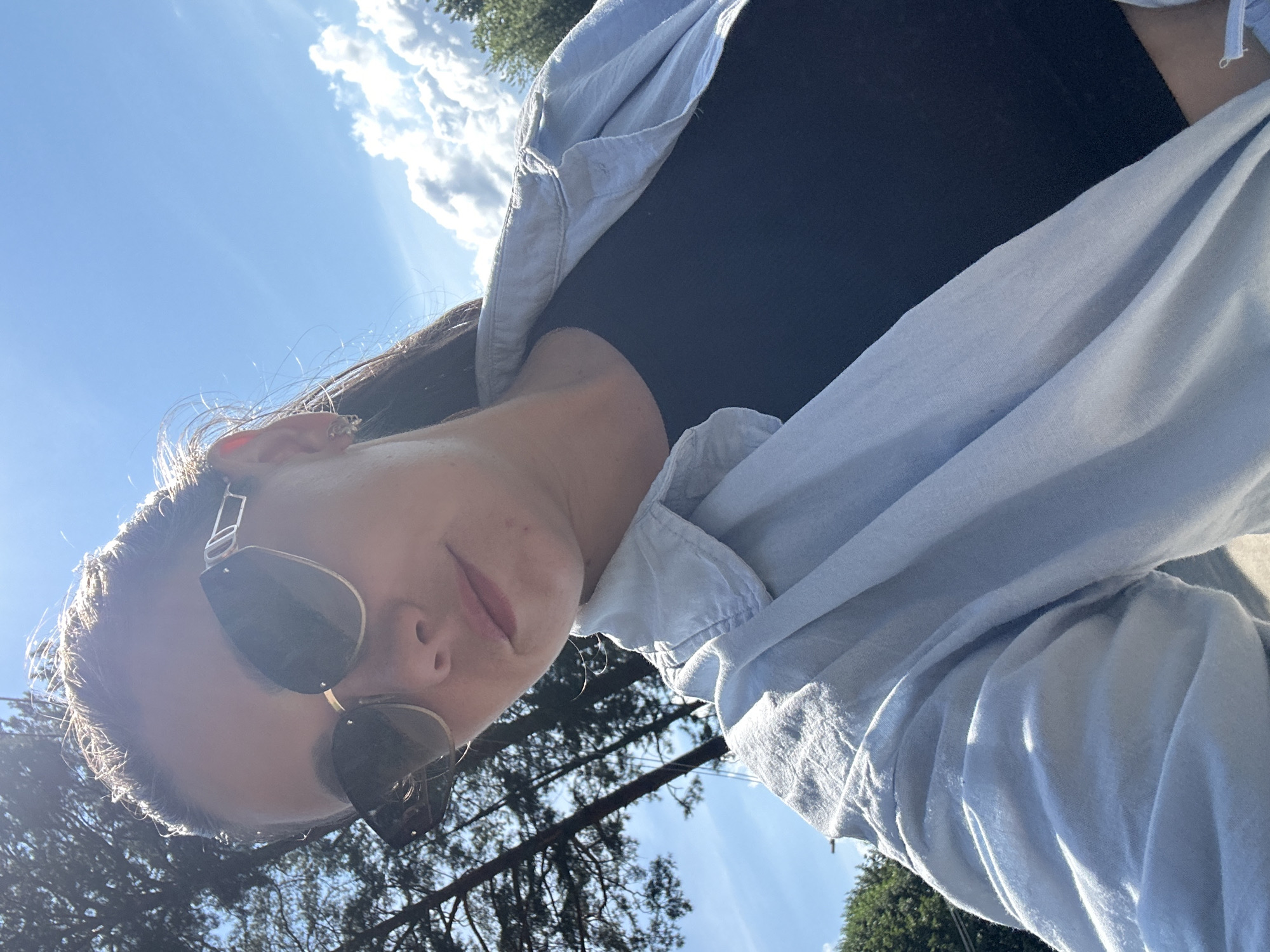A young woman is taking a selfie outdoors on a sunny day. She is wearing large, stylish sunglasses and a light blue open-collared shirt over a dark top. Her hair is dark brown and pulled back, with a few strands falling forward. The background shows a clear blue sky with some fluffy white clouds, and the tops of tall evergreen trees. The overall lighting suggests bright daylight. The scene appears to be in a natural, possibly park-like or forested area, consistent with the outskirts of Chaikovsky, Russia.