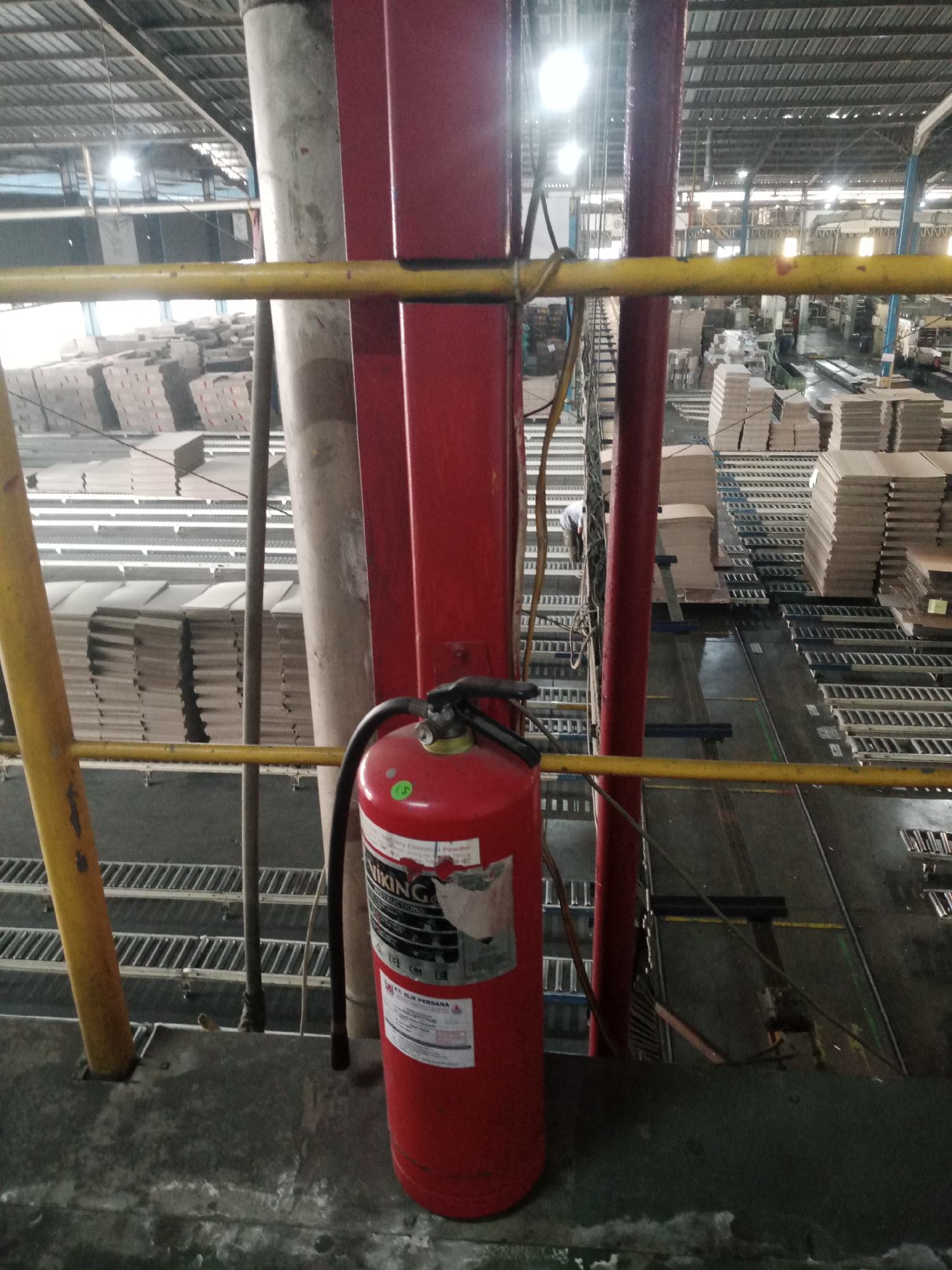 The image is an overhead view of a factory floor in Balaraja, Indonesia. The scene is industrial, filled with stacks of cardboard boxes on conveyor belts. Prominently in the foreground is a red fire extinguisher. Yellow and red metal railings and large red support columns are visible, suggesting a mezzanine or elevated walkway. Natural and artificial lights illuminate the space, creating a bright, somewhat harsh environment. The overall impression is one of a busy, functional manufacturing or warehousing facility.