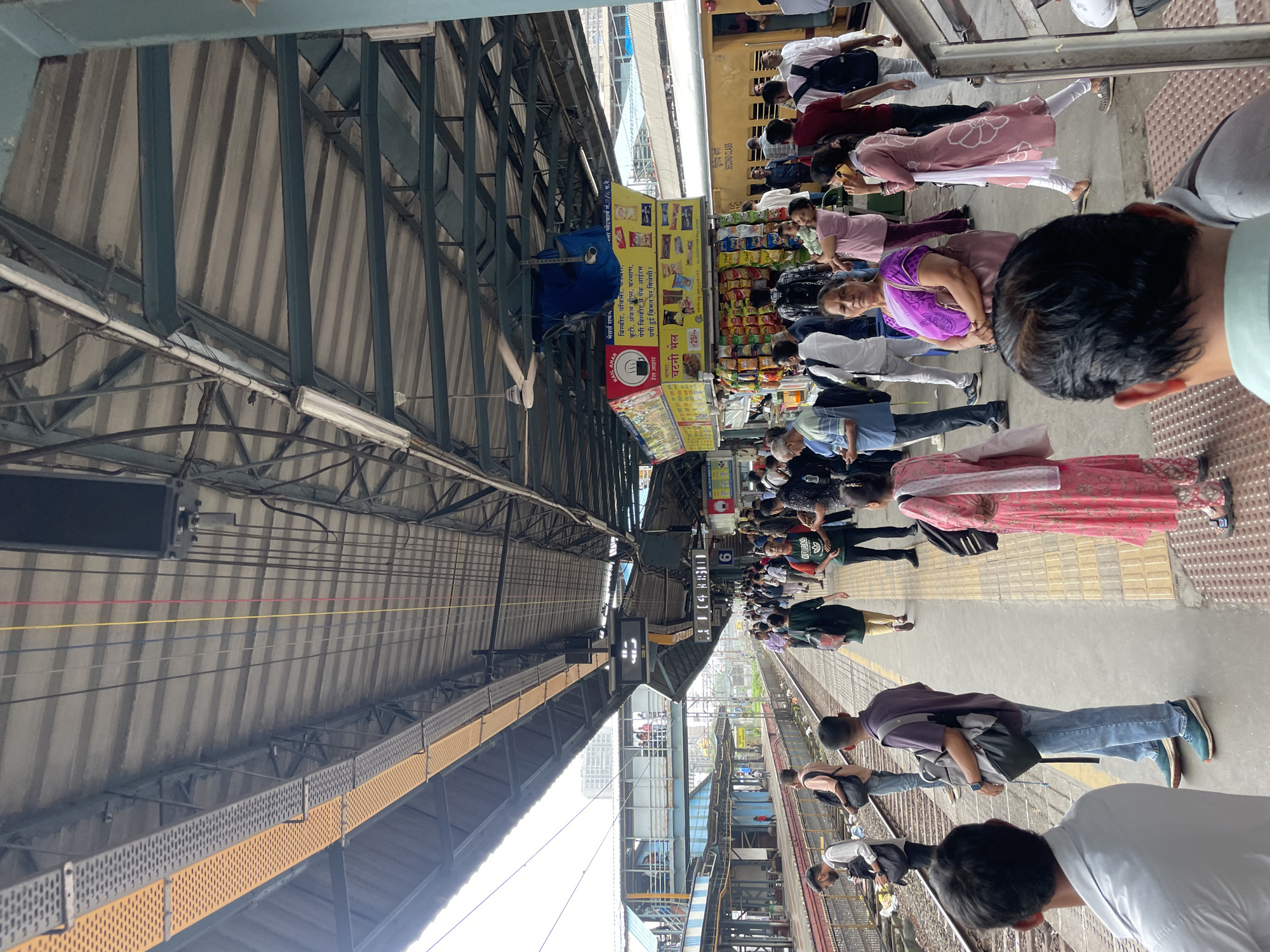 The image shows a crowded train station platform in Mumbai, India. The platform is covered by a large, corrugated metal roof supported by a dark green metal structure. Overhead, there are electrical wires and a ceiling fan. A yellow train is visible on the tracks to the right of the frame.
Numerous people are standing on the platform, many carrying bags and luggage, suggesting they are travelers. They are of various ages and genders, dressed in casual clothing, including traditional Indian attire like salwar kameez and modern Western wear. Some are looking at their phones, others are talking to each other, and some are standing still, seemingly waiting. One person in the foreground is looking away from the camera. A kiosk selling snacks is visible in the middle of the platform, with people browsing its offerings.
The platform itself is made of concrete with yellow and black textured safety markings along the edge. There are train tracks running alongside the platform. In the background, there are more train platforms and station buildings, with some higher-rise structures visible in the distance, indicating an urban environment. The overall atmosphere appears to be one of busy, everyday activity at a major transportation hub.