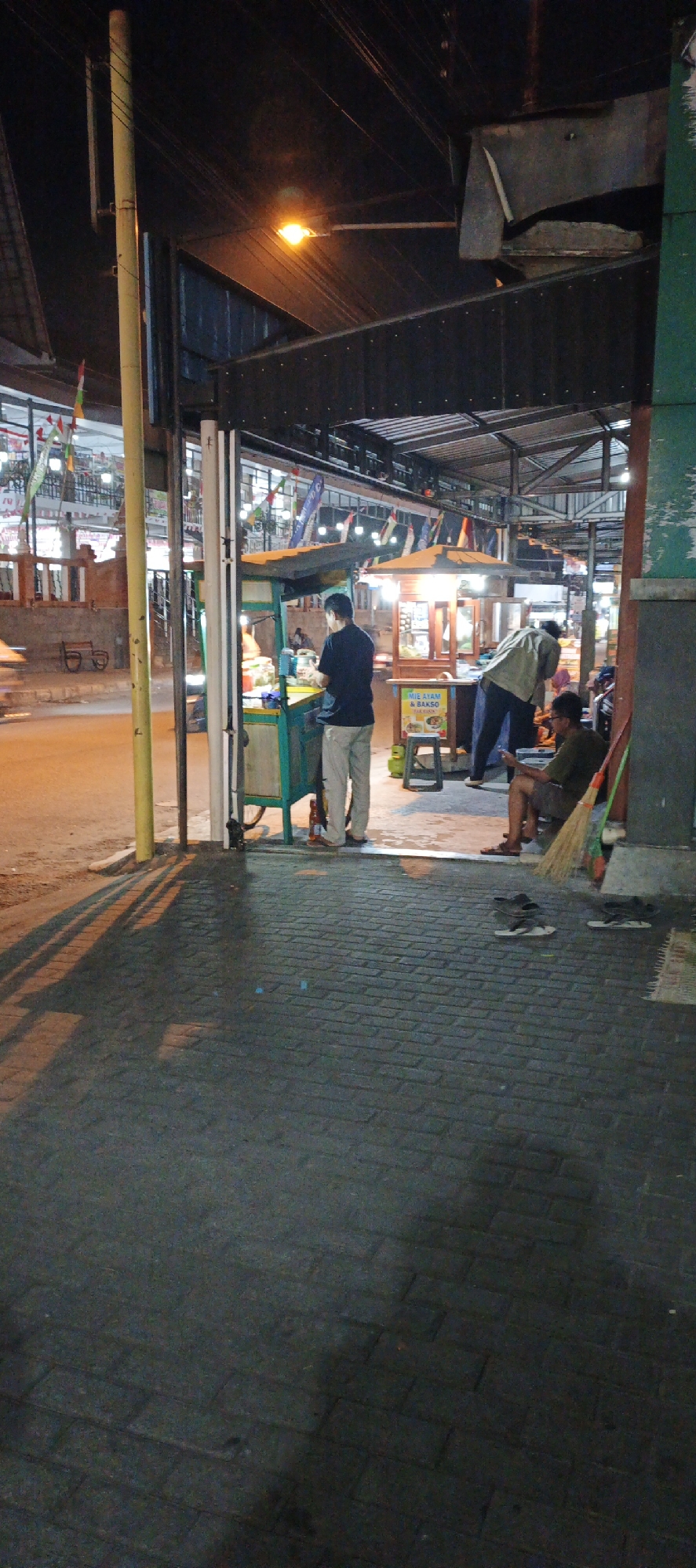 This is a nighttime photograph taken from a low angle, showing a street scene in Sidoagung, Indonesia. The foreground is a dark, paved walkway with visible brick-like patterns and shadows cast from overhead structures. In the midground, an open-air food stall is visible, illuminated by overhead lights. A man in a dark shirt and light-colored pants stands behind the counter, appearing to be serving or preparing food. Next to the stall, a menu board is displayed, featuring Indonesian text and images of food, indicating it is a food vendor.
Further into the scene, another food stall with a lighted display case is present. A person in a light-colored jacket is bending over at this stall. To the right, a man wearing a dark shirt and shorts is seated on a low stool, holding a mobile phone. A broom leans against a green vertical structure next to him. In the background, there are buildings with multiple levels, adorned with flags and string lights, suggesting a commercial or market area. The overall atmosphere is that of a bustling night market or street food area.