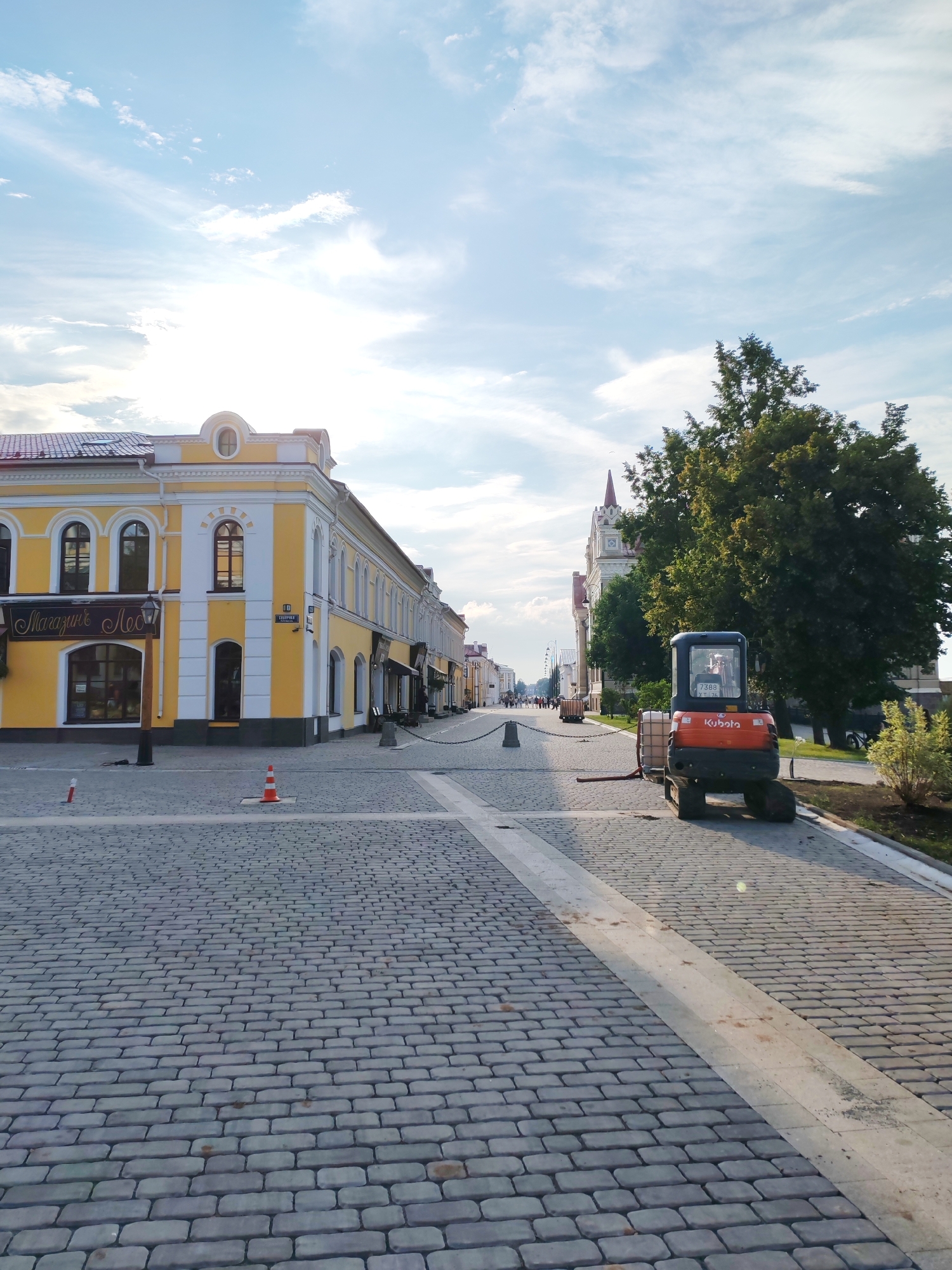 A low-angle, medium shot captures a European-style street scene in the city of Bol'shie Mkhi, Russia, under a bright, partly cloudy sky. The foreground is dominated by a cobblestone street. To the left, a building with a yellow facade and white trim features arched windows and signs. A lamppost stands in front of the building. Orange traffic cones are visible on the street. In the mid-ground, a small orange excavator, identified by the "Kubota" logo, is parked on the right side of the street. It is attached to two large white cylindrical tanks. A lush green tree stands behind the excavator. Further down the street, more buildings line the right side, and a chain barrier separates the sidewalk from the street. People are visible walking in the distance. The overall impression is one of urban development or renovation.