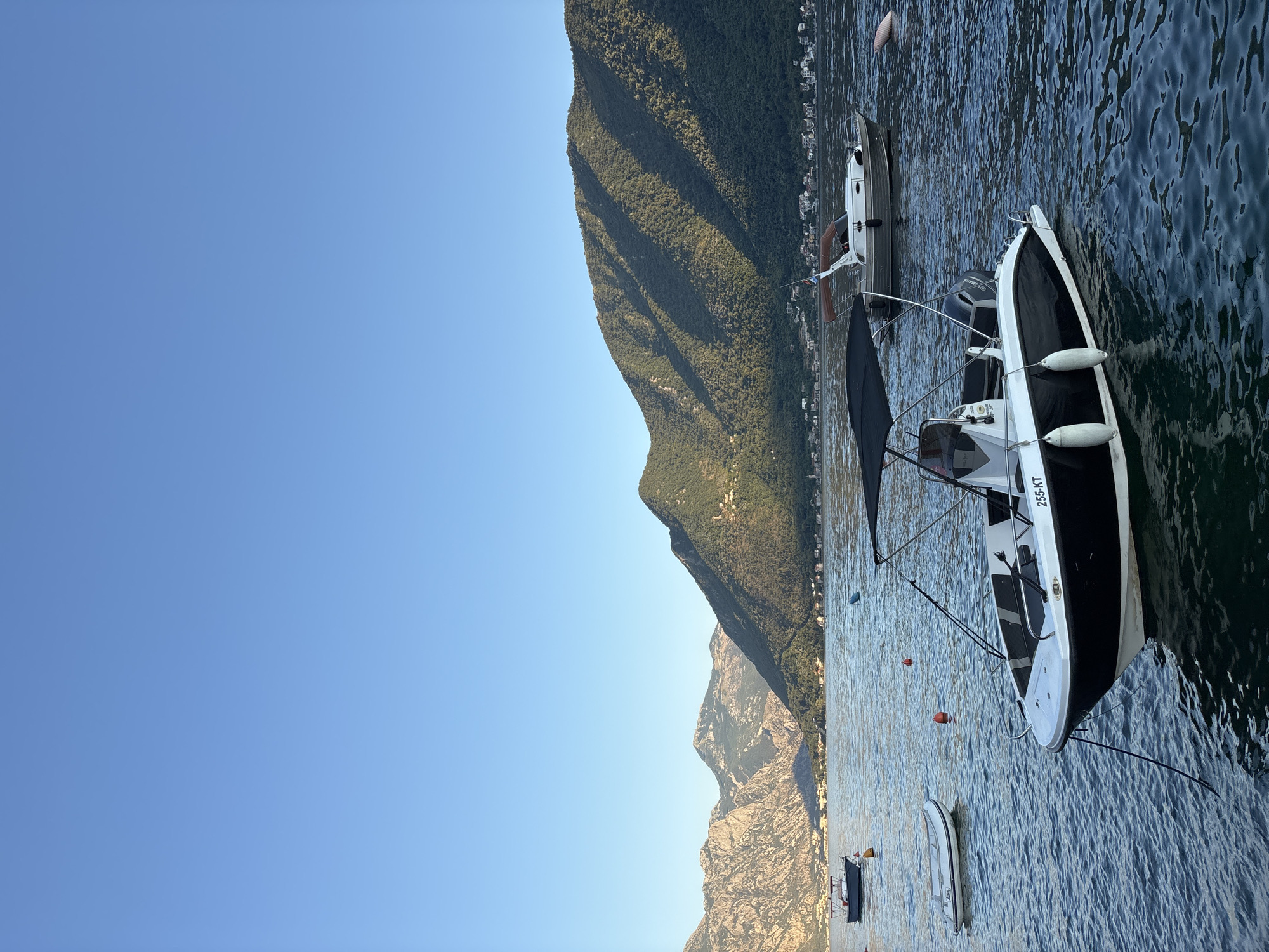 A wide-angle shot captures a serene bay near Perast, Montenegro, under a clear blue sky. Several boats are moored in the calm, dark blue water. In the foreground, a black and white speedboat with a dark canopy is anchored. Its registration number, "255-KT," is visible. Two white fenders hang from its side. Further out, a smaller white inflatable dinghy rests on the water to the left. In the mid-ground, a dark-hulled boat with a white canopy is anchored, and another small, dark boat is visible in the distance. Red buoys dot the water.
The background is dominated by steep, green, tree-covered mountains that rise dramatically from the water's edge. The sunlight casts shadows across the mountain slopes, highlighting their rugged texture. Along the base of the mountains, a scattering of buildings indicates a coastal settlement. The overall impression is one of peaceful coexistence between nature and human activity.