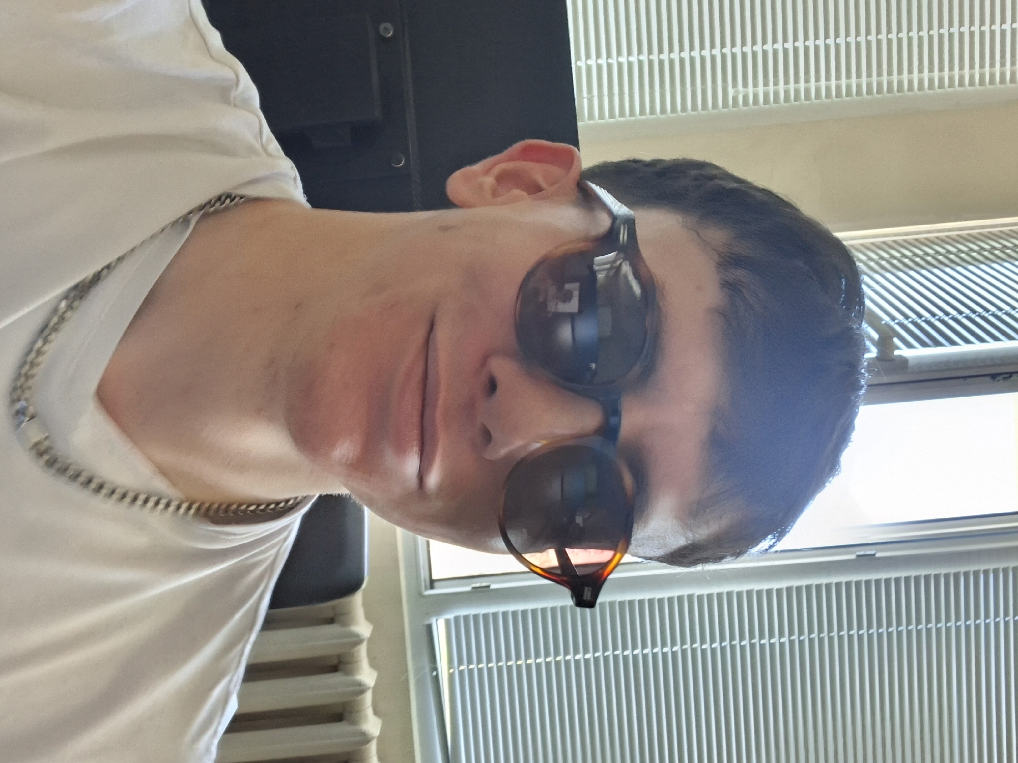 A person wearing sunglasses and a silver chain necklace is in front of a window with a white radiator. The location cannot be determined from the image.