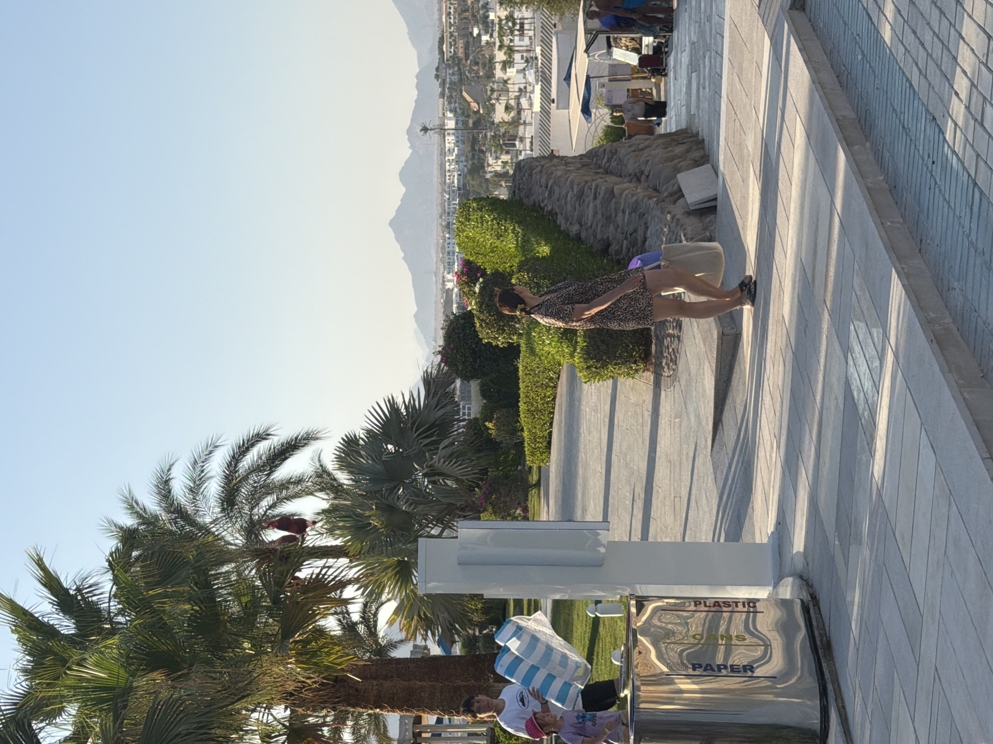 The image depicts a sunny day scene with a person standing near a recycling bin. The person, a woman wearing a patterned dress and carrying a bag, is facing away from the camera. Behind her, a white recycling bin has labels for "PLASTIC," "CANS," and "PAPER." A palm tree is visible to the left, and in the background, a city with buildings and mountains can be seen under a clear blue sky. The time of day appears to be late afternoon or early evening, as indicated by the long shadows cast by the sun. The setting is a resort or public area, likely in Sharm El Sheikh, Egypt, given the presence of palm trees and the architectural style of the buildings in the distance. The overall mood is relaxed and casual, suggesting a vacation or leisure setting.