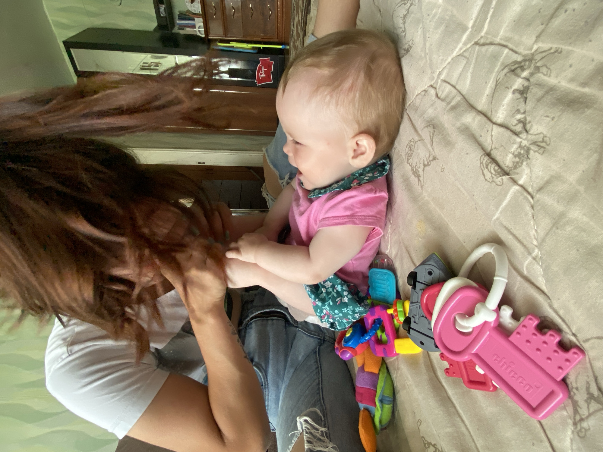 A woman with brown hair and a white shirt is holding a baby wearing a pink shirt with a floral print collar and denim shorts. The baby is lying on a bed next to a colorful toy and a set of pink and gray keys. The bed has a light-colored patterned bedspread. A dresser and other furniture are visible in the background. There is no verifiable evidence that this image was taken in the city of Фоки, Russia.