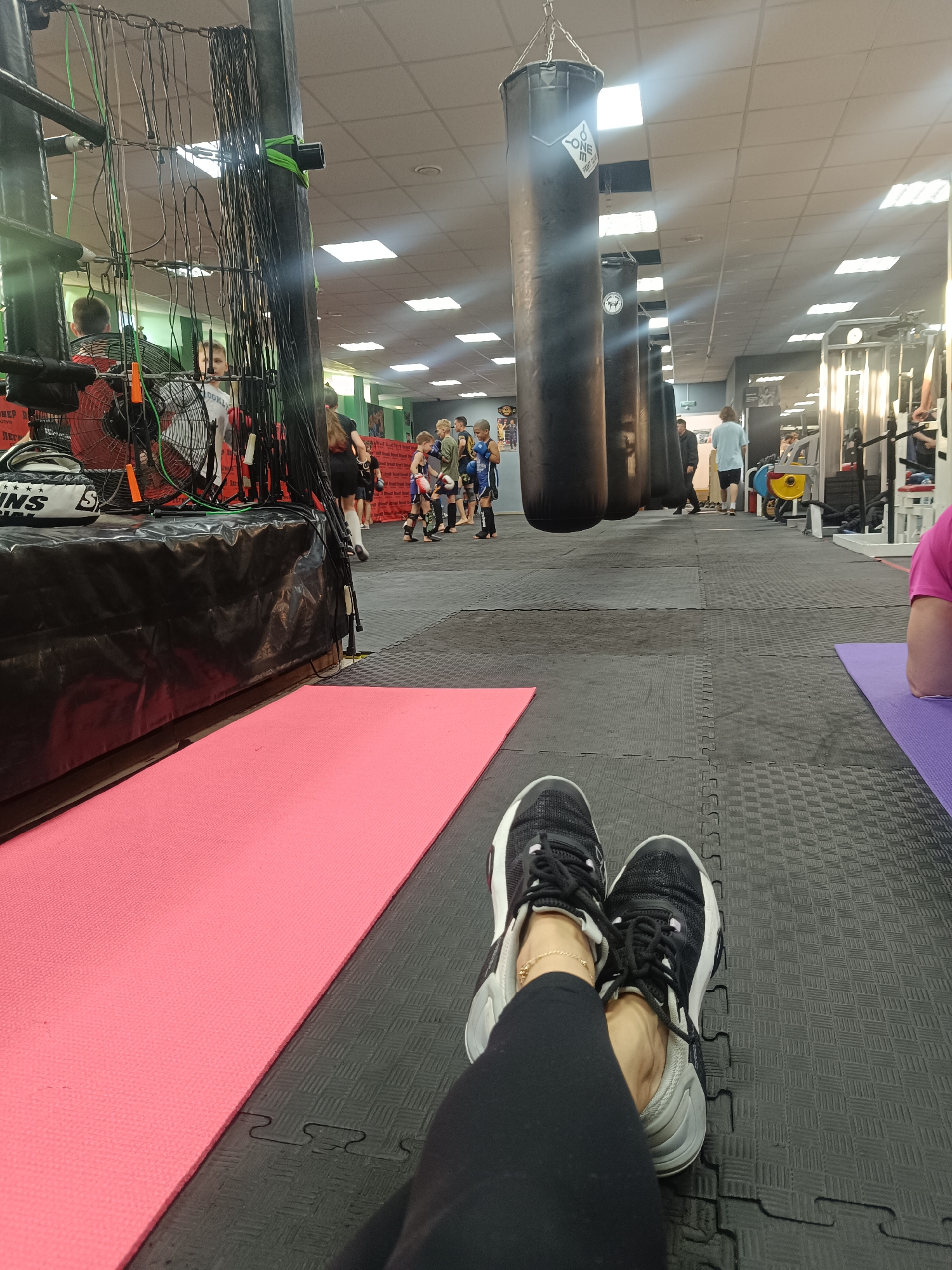 The image depicts a boxing gym in Naberezhnye Chelny, Russia. In the foreground, a person wearing black leggings and black and white athletic shoes is reclining on the floor. A pink yoga mat is to the left of the person's legs, while a purple yoga mat is to the right. Behind the person, several black punching bags hang from the ceiling. In the background, there is a boxing ring, exercise equipment, and several people, including children wearing boxing gear. Overhead fluorescent lights illuminate the gym, which has a black puzzle mat floor.