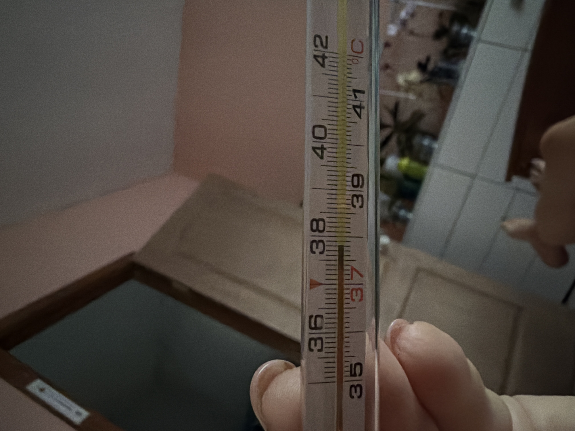 A close-up, eye-level shot captures a mercury thermometer being held by a person's fingers in what appears to be a room with a pale pink wall in the background. The thermometer displays a temperature reading between 37 and 38 degrees Celsius, with the mercury line reaching approximately the middle point between these two markings. The thermometer itself is white with black and red markings. The background includes a glimpse of a doorway and a tiled surface that seems to be a countertop or shelf, with various objects that are out of focus. The image is grainy. There is no indication of the location in the city of Chaikovsky, Russia.