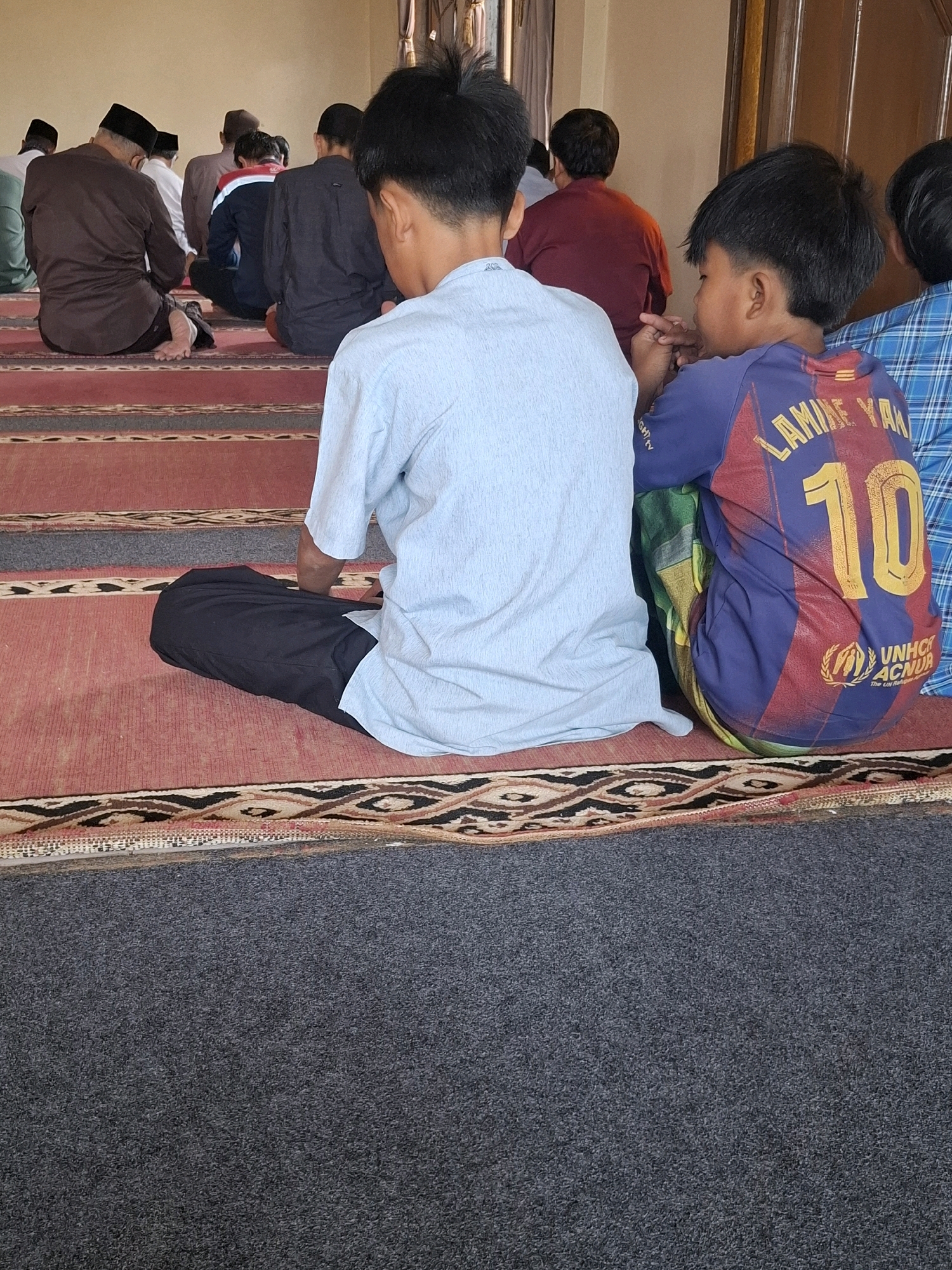 The image depicts an indoor scene, likely a prayer room, with several individuals, primarily males, seated on a carpeted floor. In the foreground, two young boys are visible from behind. The boy on the right wears a blue and red soccer jersey with the name "LAMINE YAN" and the number "10" prominently displayed on the back, along with a UNHCR logo. He is seated next to another boy wearing a light blue shirt. Several other individuals are seated further back, facing away from the camera, some wearing traditional Muslim prayer caps. The floor is covered with a red and patterned carpet. The background reveals a wall and a doorway with wooden doors. The overall setting suggests a communal gathering for prayer or religious instruction. The location is identified as Ciheulang Tonggoh, Indonesia.