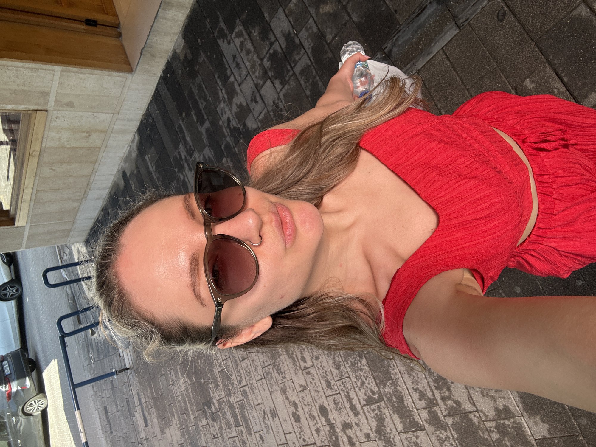 A woman in a red dress and sunglasses is positioned in an outdoor setting on what appears to be a paved walkway. She's holding a bottle of water. In the background, there is a street with parked cars and a blue bicycle rack. There's also a building with a wooden door. Given the prompt, this scene is located in the city of Fethiye, Turkey.