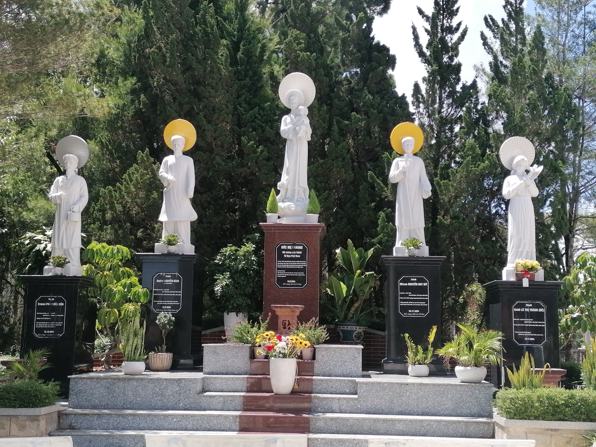 The image showcases a series of five white religious statues on pedestals set against a backdrop of tall evergreen trees. The central statue is of a woman holding a child, possibly the Virgin Mary. The statues to the left and right of the central figure appear to be saints or religious figures, with yellow halos or parasol-like sunshades above their heads.
Each statue stands on a black or red polished stone pedestal with inscriptions in Vietnamese. Small potted plants decorate the bases of the pedestals, and a concrete staircase leads up to the display. The location is implied to be in the city of Da Lat, Vietnam.