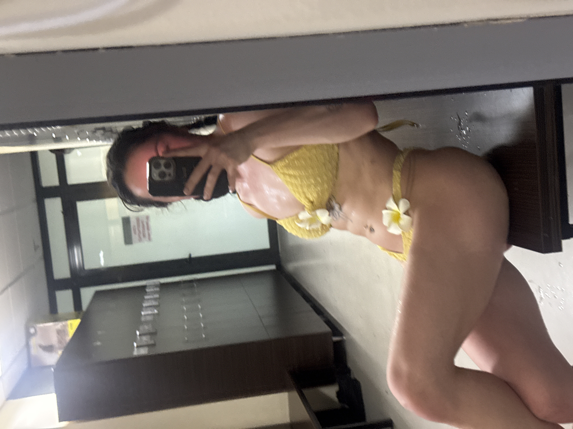 A woman is reflected in a mirror while taking a selfie with a smartphone. She is wearing a yellow bikini with white flower embellishments. The background shows a locker room with rows of lockers and a doorway. The location is possibly a gym or changing room in Burgas, Bulgaria.