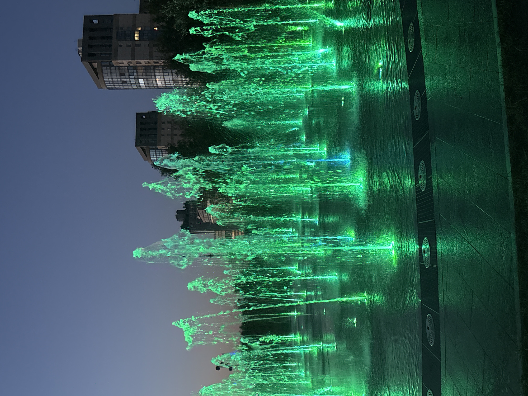 The image depicts a nighttime scene of a vibrant, illuminated fountain in Saint Petersburg, Russia. The fountain's water jets are lit with a vivid green glow, creating a dynamic and visually striking display. In the background, modern high-rise buildings are visible against the darkening twilight sky, suggesting an urban setting. The foreground shows the paved area surrounding the fountain, with some greenery at the very bottom edge of the frame. The scene is devoid of any people or animals, focusing solely on the architectural and natural elements of the park or public space.