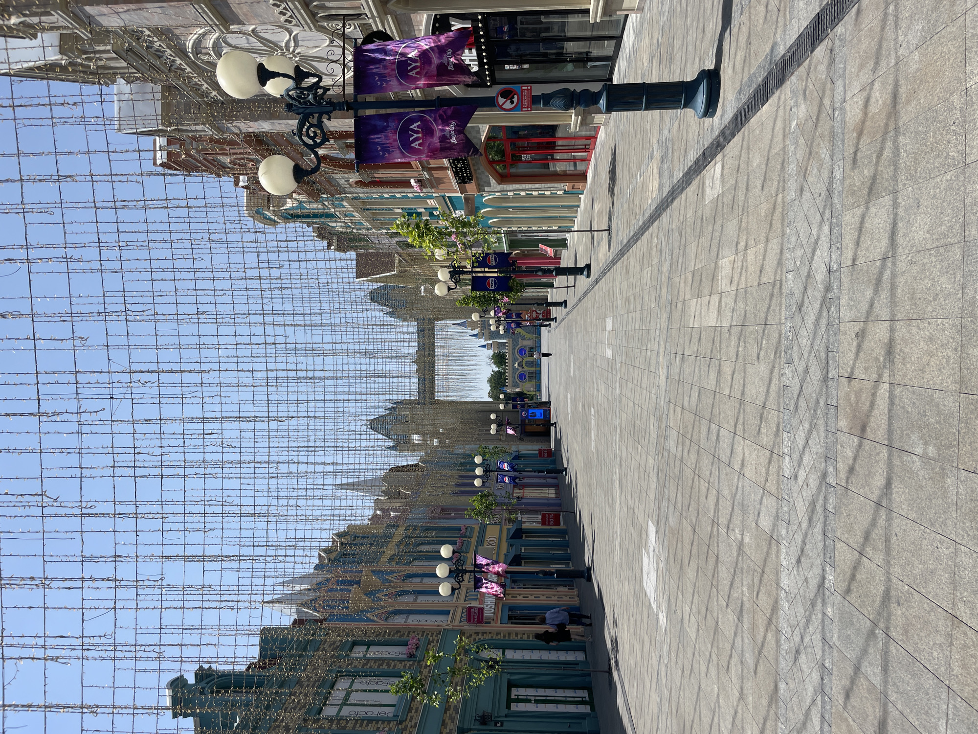 This is a wide shot of a pedestrian street lined with colorful buildings, possibly in a themed entertainment district, under a canopy of strings of lights. The architecture is ornate, with decorative facades and arched windows. In the foreground, the street is paved with large, square tiles, and there are lampposts with multiple globe lights spaced along the walkway. A few trees are planted along the sides of the street. In the distance, a large arched structure with spires is visible, resembling a castle gate. A few people are walking on the street, and some are looking into shop windows. The sky is clear blue. The context provided indicates this location is in Tashkent, Uzbekistan.