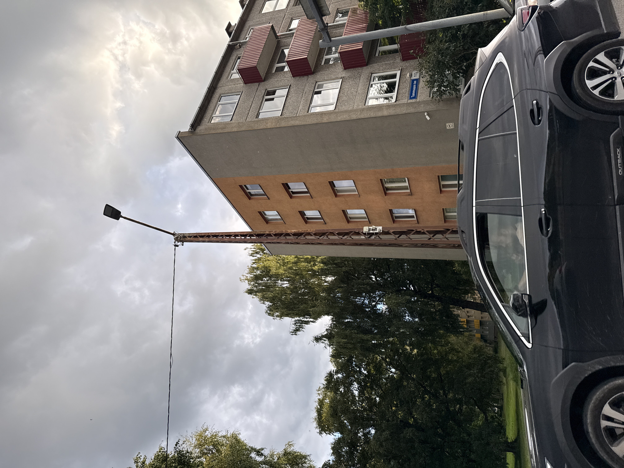 The image shows a black SUV driving on a street in Tallinn, Estonia, with a multi-story apartment building in the background. The building is a mix of grey and orange facades, with several balconies featuring red railings. A street sign on the building reads "Tammsaare tee 113". A streetlamp with a rusty metal pole stands next to the building. Trees, some with green foliage, are visible on the left and right sides of the image. The sky is overcast with clouds. A person can be seen through the SUV's window.