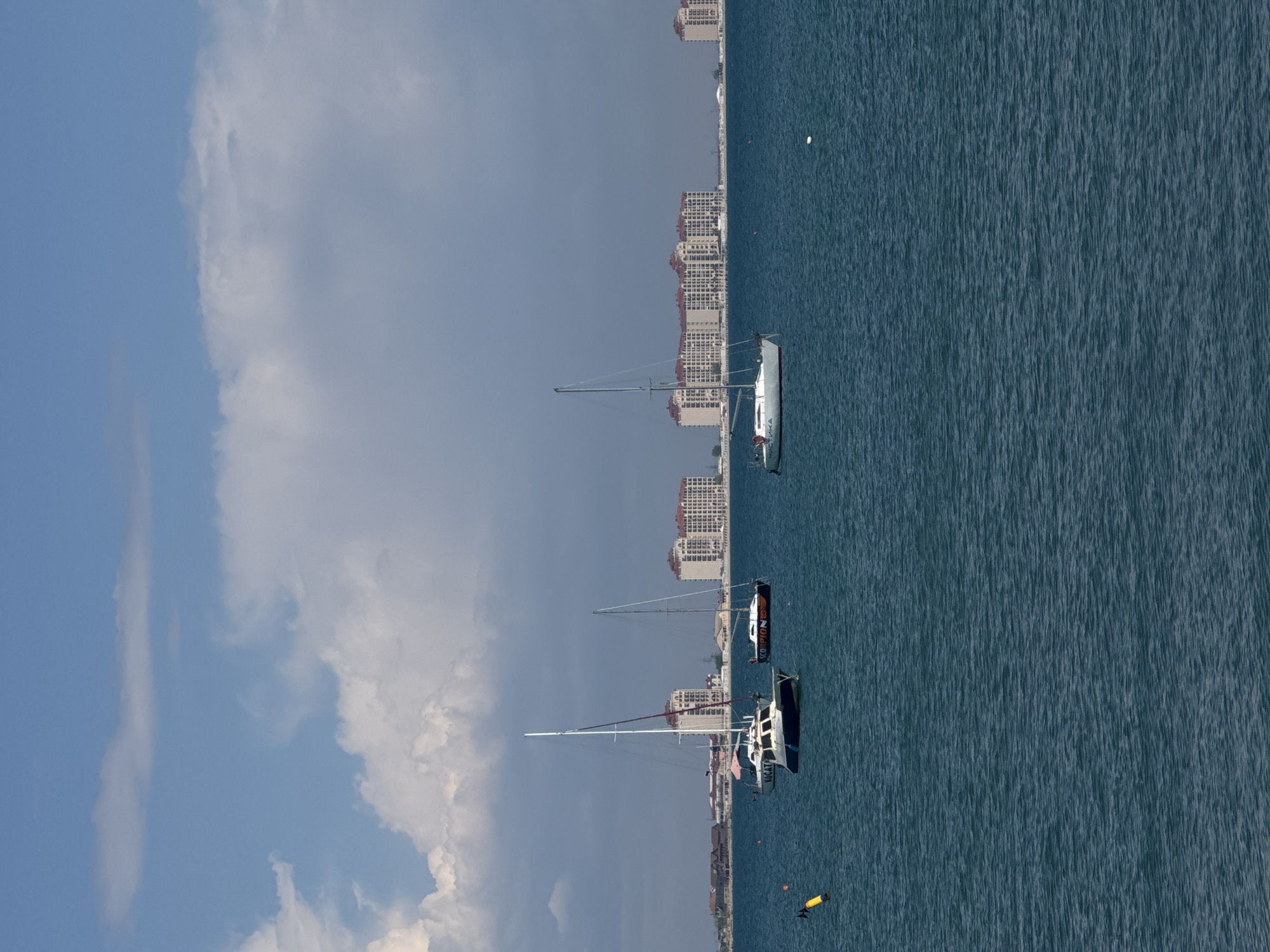 The image depicts a coastal scene, with the city of Yevpatoriya, Ukraine, in the background. The foreground features the Black Sea, in which several sailboats are anchored. The sky is partly cloudy. Buildings are visible along the coastline.
