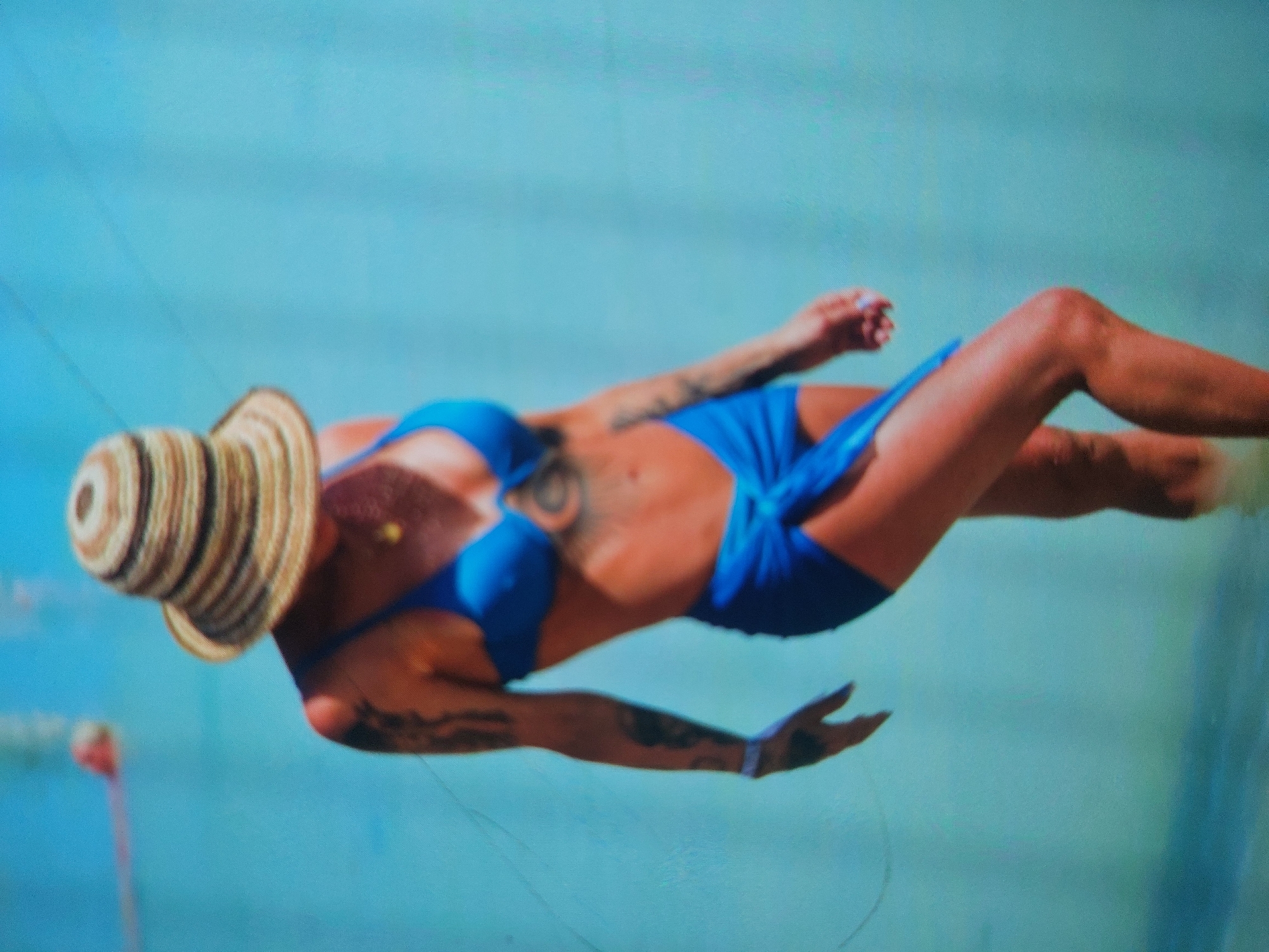 The image depicts a person, likely female, suspended horizontally against a vibrant blue background, which appears to be either the clear sky or expansive open water. The individual is wearing a bright blue two-piece bikini and a striped straw hat. They exhibit a tanned complexion and several visible tattoos on their arms and torso, including a prominent circular design on their side.
The pose and background elements strongly suggest an aerial watersport such as parasailing or kitesurfing. Faint lines, possibly ropes or lines from a kite or parachute, are visible extending into the blue backdrop. In the lower left corner, a blurry pink object with a thin line further hints at sporting equipment. The overall scene conveys a sunny and clear day, typical for watersport activities.
Given the provided location context of Tsarevo, Bulgaria, a coastal town, the activity is almost certainly taking place over the Black Sea. The image is taken during daylight hours. No specific time of day, weather details beyond "clear," or emotions are discernible. There is no visible text in the media file.
