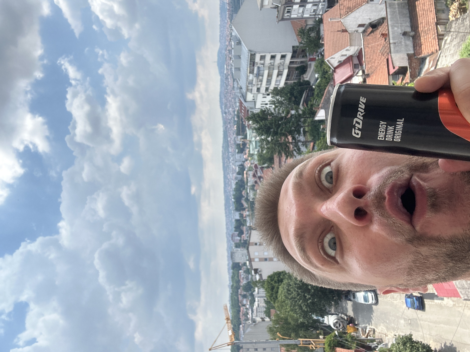 The image shows a close-up selfie of a man holding a can of "G'DRIVE ENERGY DRINK ORIGINAL". The man has a surprised expression with wide eyes and an open mouth. In the background, there is a cityscape with buildings, trees, and a crane, suggesting an urban environment. The sky is cloudy. The backdrop appears to be from Zvezdara Urban Municipality, Serbia, given the characteristic architecture of buildings and the presence of a construction crane, which are common in developing urban areas of Serbia.
