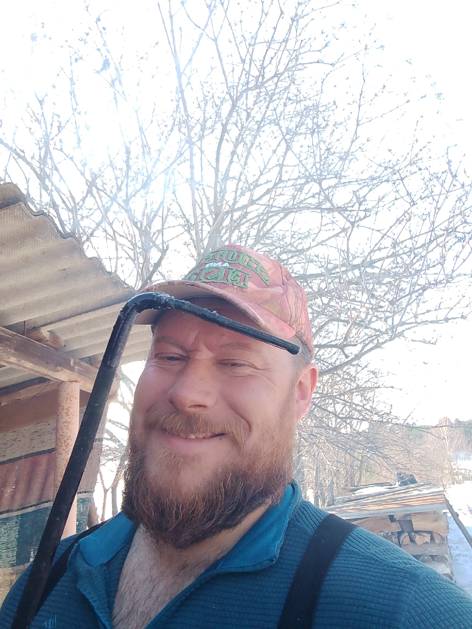 An adult male with a reddish-brown beard and mustache smiles at the camera, situated outdoors in Foki, Russia. He wears a camouflage-patterned baseball cap with green text and a teal ribbed quarter-zip shirt, with a black suspender strap visible on his right shoulder. A dark, bent metal object, possibly a tool, with light residue near the bend, is positioned horizontally across his upper face, beneath the cap. The background features a corrugated asbestos-cement roof on the left, supported by wooden beams, and numerous bare deciduous trees against a bright, possibly overcast or hazy, sky. To the right, a stack of firewood is visible, topped with a makeshift corrugated roof, with snow covering the visible ground.