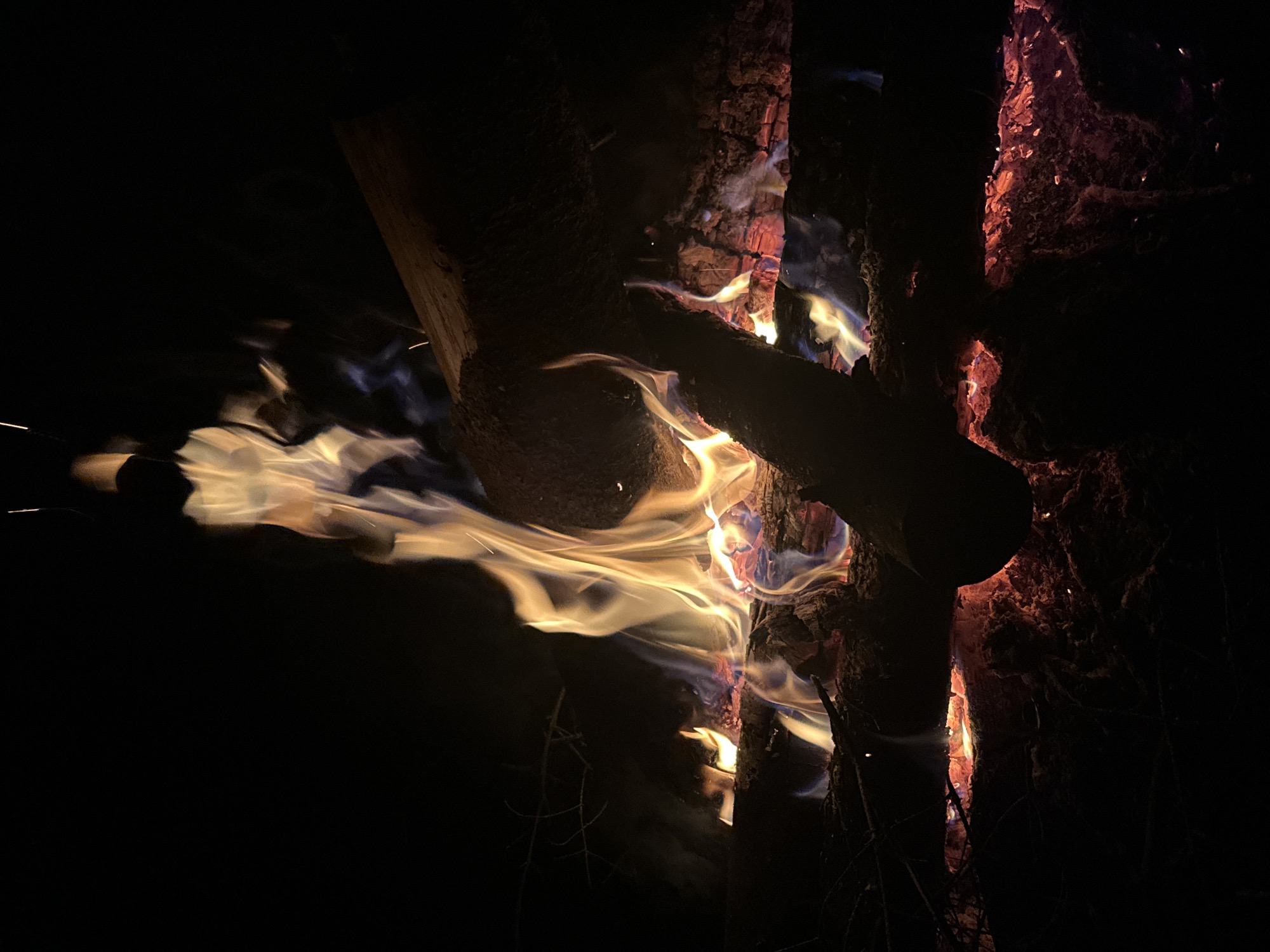 The image depicts a close-up view of a bonfire at night. The fire burns intensely between stacked logs, with flames exhibiting hues of yellow, orange, and hints of blue. The bark of the logs is charred and partially illuminated by the firelight. Sparks are visible, flying off from the blaze. The background is predominantly dark, emphasizing the intensity of the fire. The location is not determinable from the image. There are no people, explicit actions or interactions, or specific objects beyond the fire and wood. The environment is a dark, unidentifiable outdoor setting.