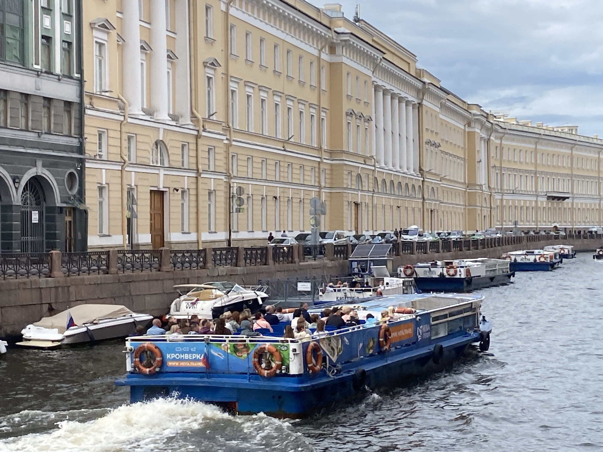 The image depicts a tour boat filled with passengers navigating a canal in Saint Petersburg, Russia. The canal runs alongside a grand, long, multi-story building characterized by a facade of cream-colored walls, numerous windows, and tall white columns. The building appears to be of historical or governmental significance. The canal water is disturbed by the wake of the tour boat, indicating movement. Several other boats are visible docked or moving in the canal, and a stone embankment lines the opposite side of the canal. Cars and people are visible on the embankment. The sky is overcast with clouds. The tour boat is predominantly blue with orange life preservers attached to its railing and signage with Cyrillic text.