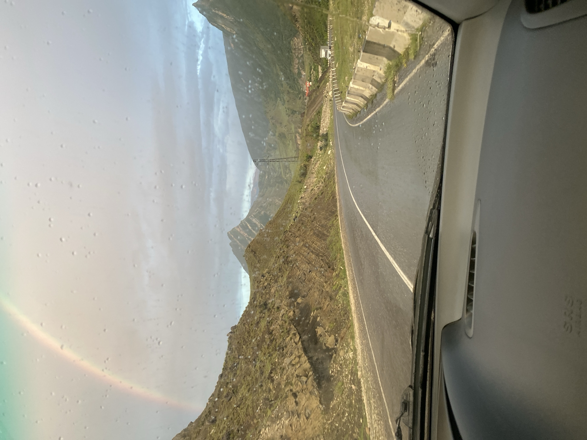 This image shows a view from inside a car driving on a wet, paved road in the mountainous region of Chalda, Russia. A faint rainbow arches across the sky, partially obscured by rain droplets on the car's windshield. The road curves to the right, with steep, rocky, and grassy slopes on the left and a guardrail along the right. In the distance, more mountains are visible, along with a power line tower and some buildings. The sky is overcast with clouds. The dashboard of the car is in the foreground.