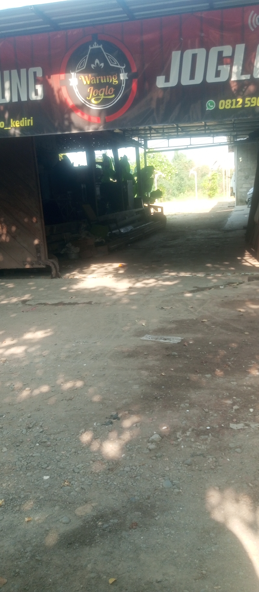 A banner displays "Warung Joglo" with a phone number "0812 590" and "o_kediri" below it. The banner is hung above an open-air structure. Inside the structure, there are various objects and machinery that appear to be metalwork or industrial equipment, partially obscured by shadows. The ground in front of the structure is covered with dirt, gravel, and fallen leaves, with dappled sunlight casting shadows. The overall scene suggests an outdoor workshop or business establishment. The location of Kediri City, Indonesia, is indicated by text on the banner.