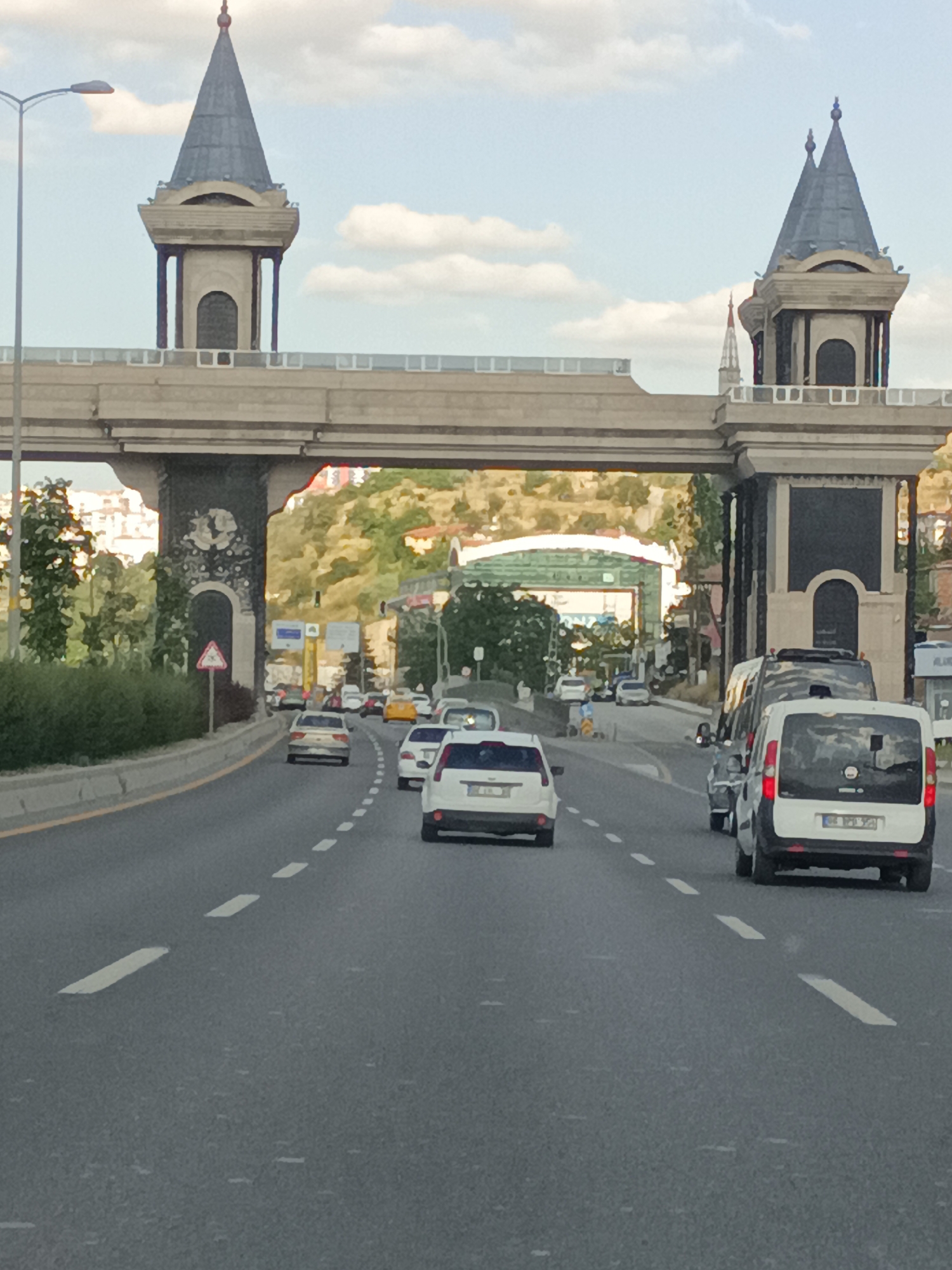 The image shows a view from a vehicle driving on a highway. The highway passes under a large, ornate gateway structure that resembles a castle. The gateway has two imposing towers with conical roofs, connected by a wide, arched bridge. The towers are decorated with columns and arched windows. The scene is set during the daytime, with a partly cloudy sky.
Several cars are driving on the highway in the same direction as the camera. These include a white van, a silver sedan, and a white hatchback. In the background, beyond the gateway, a hilly landscape with buildings and trees is visible. The weather appears to be clear and pleasant.
The gateway and the surrounding architecture suggest a location with a unique or historical theme. The presence of traffic on the highway indicates it is a functional road, likely in an urban or suburban area. The image captures a moment of transit, with the gateway serving as a prominent landmark. The overall mood is neutral, focused on documenting the visual elements of the scene. There is no visible text in the image that is legible enough to transcribe.