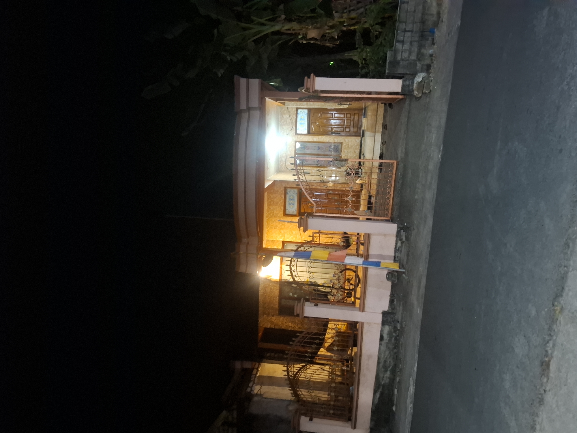 The image depicts a nighttime view of a residential property in Bulubrangsi, Indonesia. The house is illuminated by exterior lights, casting a warm glow on its facade. The house features a light-colored, textured exterior wall and a prominent tiled roof. An ornate metal gate guards the entrance to the property, with a similar fence running along the front. To the right of the house, there are several large, leafy trees. A paved road runs in front of the property, and the scene is captured at night, with the sky appearing dark. There are no people visible in the image.