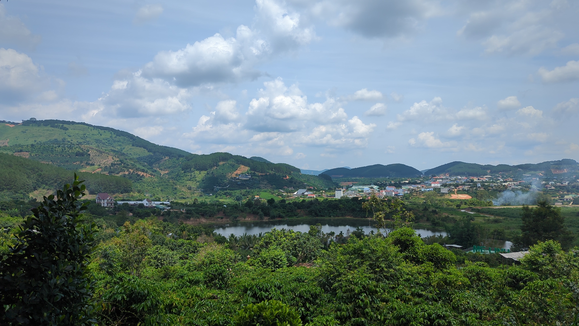 This is a wide-angle, eye-level shot of a valley with a town and surrounding hills. In the foreground, a dense canopy of lush green trees, possibly coffee plants due to the leaf shape, dominates the lower third of the frame. A large, dark-leafed tree is prominent on the left.
Beyond the trees, a calm, dark body of water, likely a lake or reservoir, occupies the middle ground. Several structures are visible on its banks, including what appears to be a large, multi-story building with a reddish-brown roof on the left side, and smaller, white houses scattered among the greenery.
The mid-ground also features a town with numerous buildings, predominantly white and reddish-orange roofs, spreading across the valley floor and up the gentle slopes of the hills. Smoke plumes rise from a few locations within the town, suggesting burning or industrial activity. Some agricultural terracing is visible on the distant hillsides.
The background consists of rolling, verdant hills that recede into the distance. The hills are covered in a mix of trees and vegetation, with some patches of lighter soil indicating human activity.
The sky above is a vibrant blue, dotted with numerous puffy, white cumulus clouds, some of which cast shadows on the landscape. The lighting suggests a bright, possibly midday, sun. The overall impression is a rural landscape with a developed settlement nestled within the hills. The location is identified as Thôn 5, Vietnam.