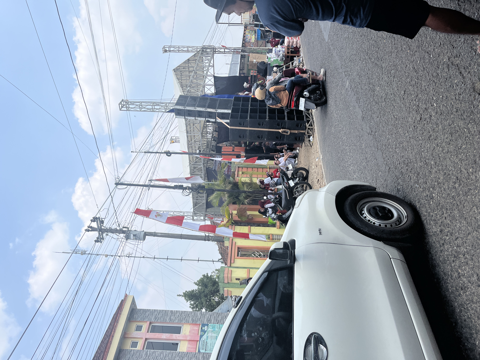 The image depicts an outdoor scene on a sunny day, likely in the city of Glagah Kulon, Indonesia, with a clear blue sky and scattered clouds. In the foreground, a white car is partially visible on the left side of the frame, with its front driver's side door and wheel in view. The car is parked on an asphalt road.
The background reveals a street scene that appears to be set up for an event. A large stage structure with scaffolding and a white roof is prominent, suggesting a performance or gathering. Piles of large black speaker cabinets are stacked high in front of the stage. Several people are gathered in the area, some sitting on the ground, others standing, and a few are on motorcycles. Indonesian flags, red and white, are displayed on poles, indicating a local celebration or event. A utility pole with numerous wires runs across the sky, a common sight in many urban and semi-urban areas. The surrounding environment includes buildings with colorful facades and some vegetation, including palm trees.
On the right side of the road, a person wearing a dark blue shirt and black shorts is standing, looking towards the event. A motorcycle with two people riding it is moving away from the camera on the right side of the road, in front of the speaker stacks. The overall impression is of a lively outdoor event taking place in a community setting.
