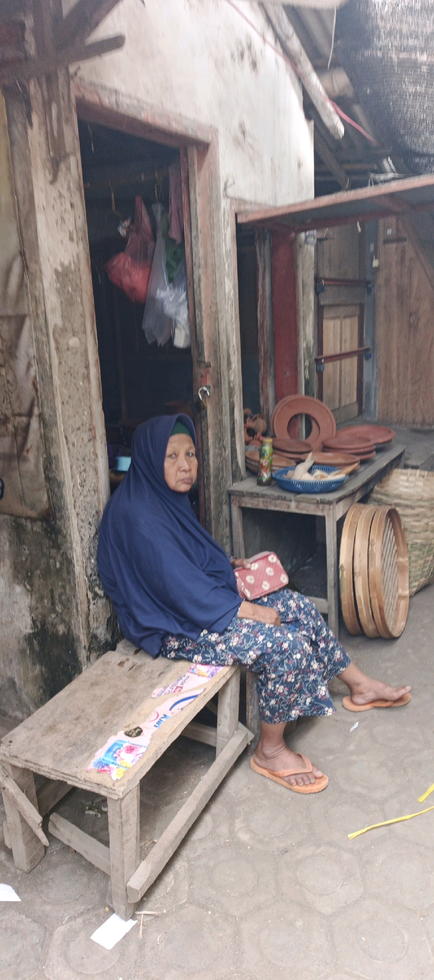 An elderly Indonesian woman wearing a dark blue hijab and a floral-patterned dress sits on a wooden bench outside a rustic building. The building appears to be made of weathered wood and concrete, with an open doorway revealing hanging garments. To her right, a wooden table displays several clay plates, a blue basket containing raw chicken, and a bottle of what appears to be a beverage. Behind the table, a large woven basket is visible, along with part of a wooden structure. The ground is paved with hexagonal tiles, and scattered paper debris is present. The overall impression is of a scene in a humble, possibly rural or semi-urban, outdoor market or residential area in Kasiyan, Indonesia.