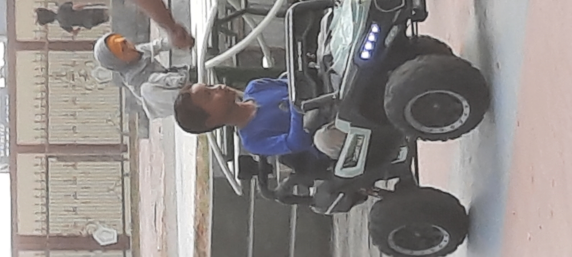 In Kanigoro, Indonesia, a young boy with dark hair and brown eyes is sitting in the driver's seat of a black toy ATV. He is wearing a long-sleeved blue shirt. The ATV has large, rugged tires and visible headlights. In the background, another child wearing a grey hooded garment and an orange headscarf is crouched down. Further back, a silhouette of another child in a dark t-shirt is standing. The scene appears to be outdoors, with a metal fence and a light-colored wall in the background.