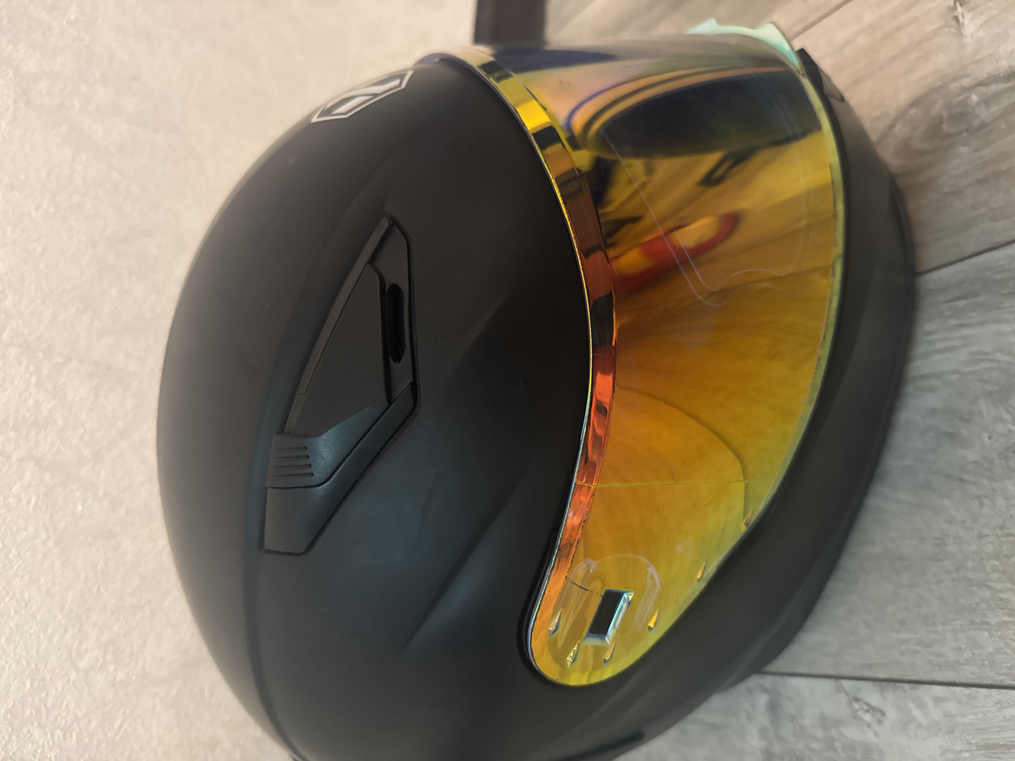 A black motorcycle helmet with a reflective gold visor is resting on a light-colored wooden floor, with a textured off-white wall in the background. The helmet has a vent on the top and a white logo on the right side. The visor shows reflections of what appear to be interior lights and possibly a red object. The image is a close-up shot, focusing on the front and side of the helmet. No people or specific city landmarks are visible.