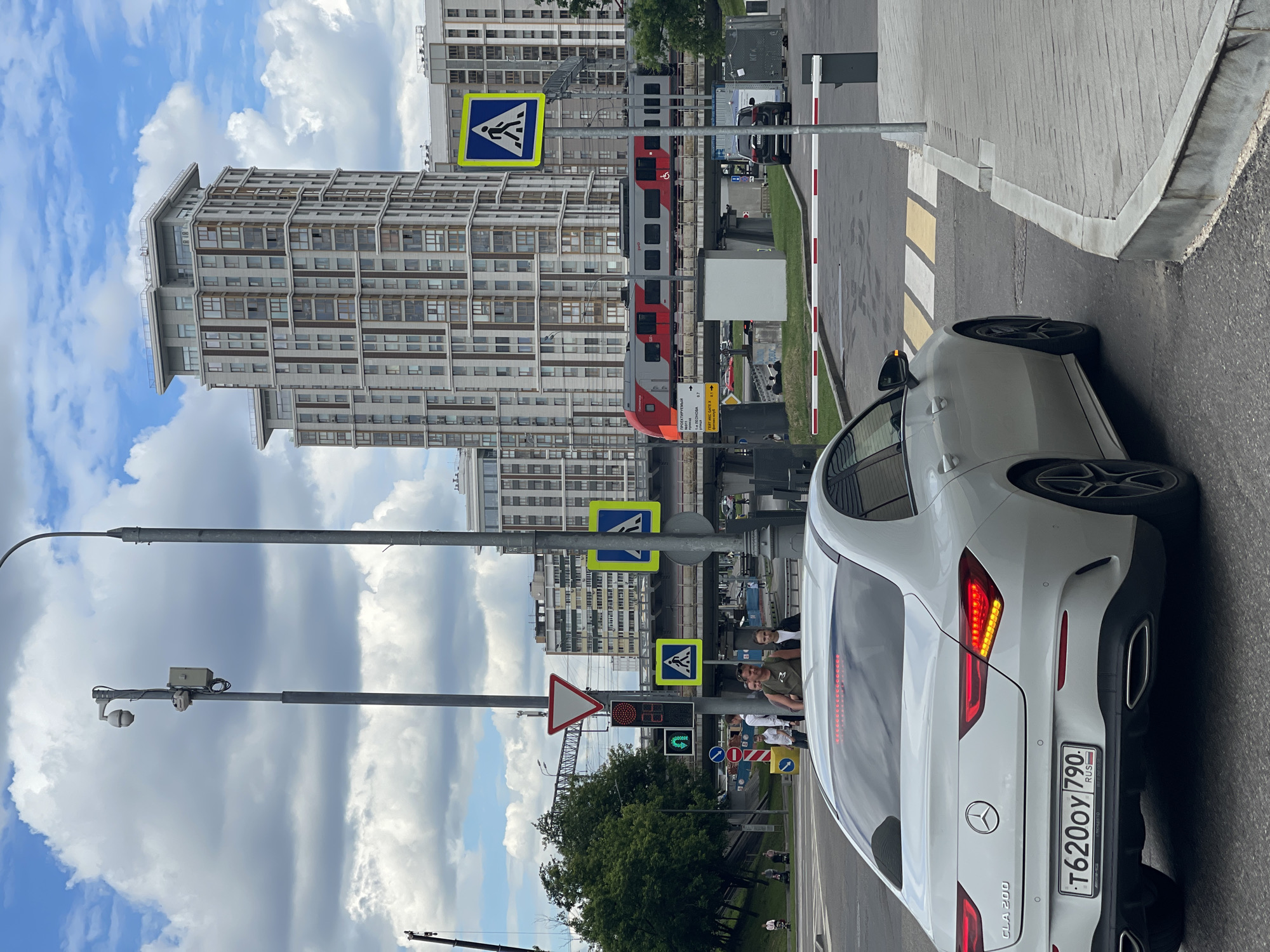A white Mercedes-Benz CLA 200 is parked at a crosswalk with a red and white barrier gate in the background. The car's license plate is visible and reads "T6200Y 790". In the background, a red and gray train is passing on an elevated railway track. A modern high-rise building stands behind the train tracks. Several pedestrians are visible near a traffic light and pedestrian crossing signs. The sky is cloudy with patches of blue. The scene appears to be an urban environment.