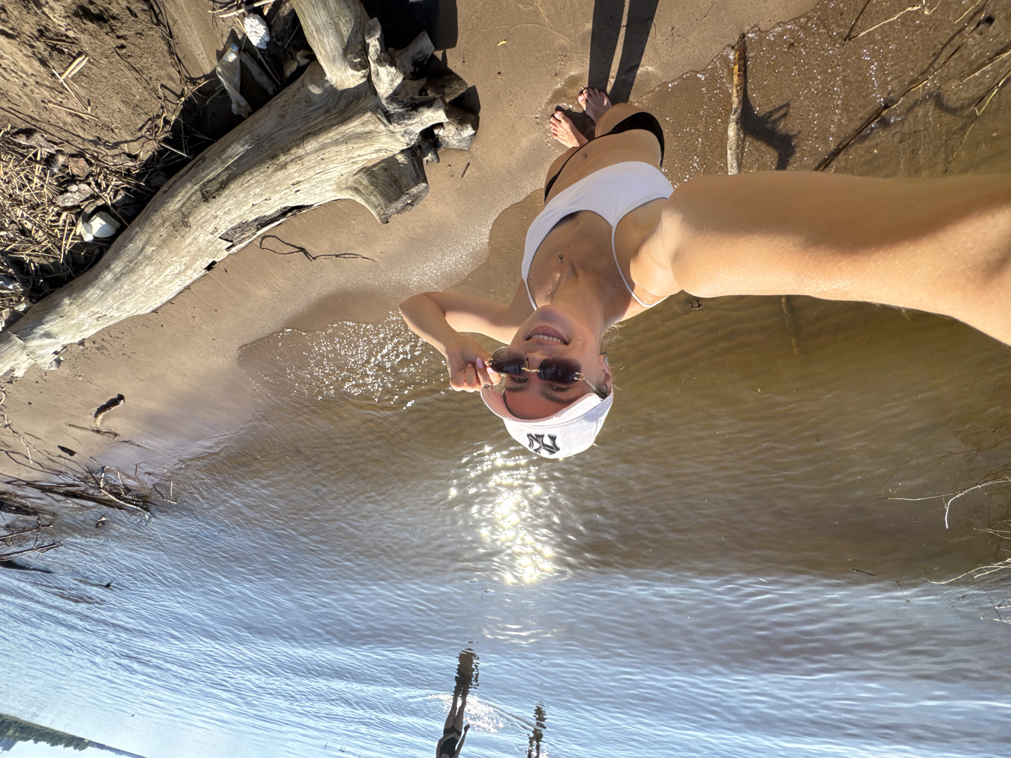 This is a selfie taken by a woman on a sunny day. She is standing on a sandy shore next to a body of water. The woman is wearing a white bikini top, black bottoms, and a white baseball cap with a New York Yankees logo. She is also wearing sunglasses and is holding a pair of sunglasses in her hand. The background shows a large piece of driftwood on the shore and the water, which appears to be a river or lake. The sun is shining brightly, reflecting off the water. The image is oriented such that the woman appears to be upside down.