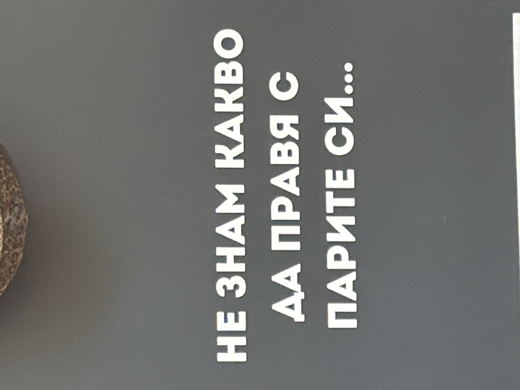 The image shows a close-up of a sign with Cyrillic text in white against a gray background. The text reads "НЕ ЗНАМ КАКВО ДА ПРАВЯ С ПАРИТЕ СИ...". The sign appears to be outdoors, possibly located in Sofia, Bulgaria given the language. There is a portion of an object, that is difficult to identify due to the angle and crop of the image, in the left side of the frame.