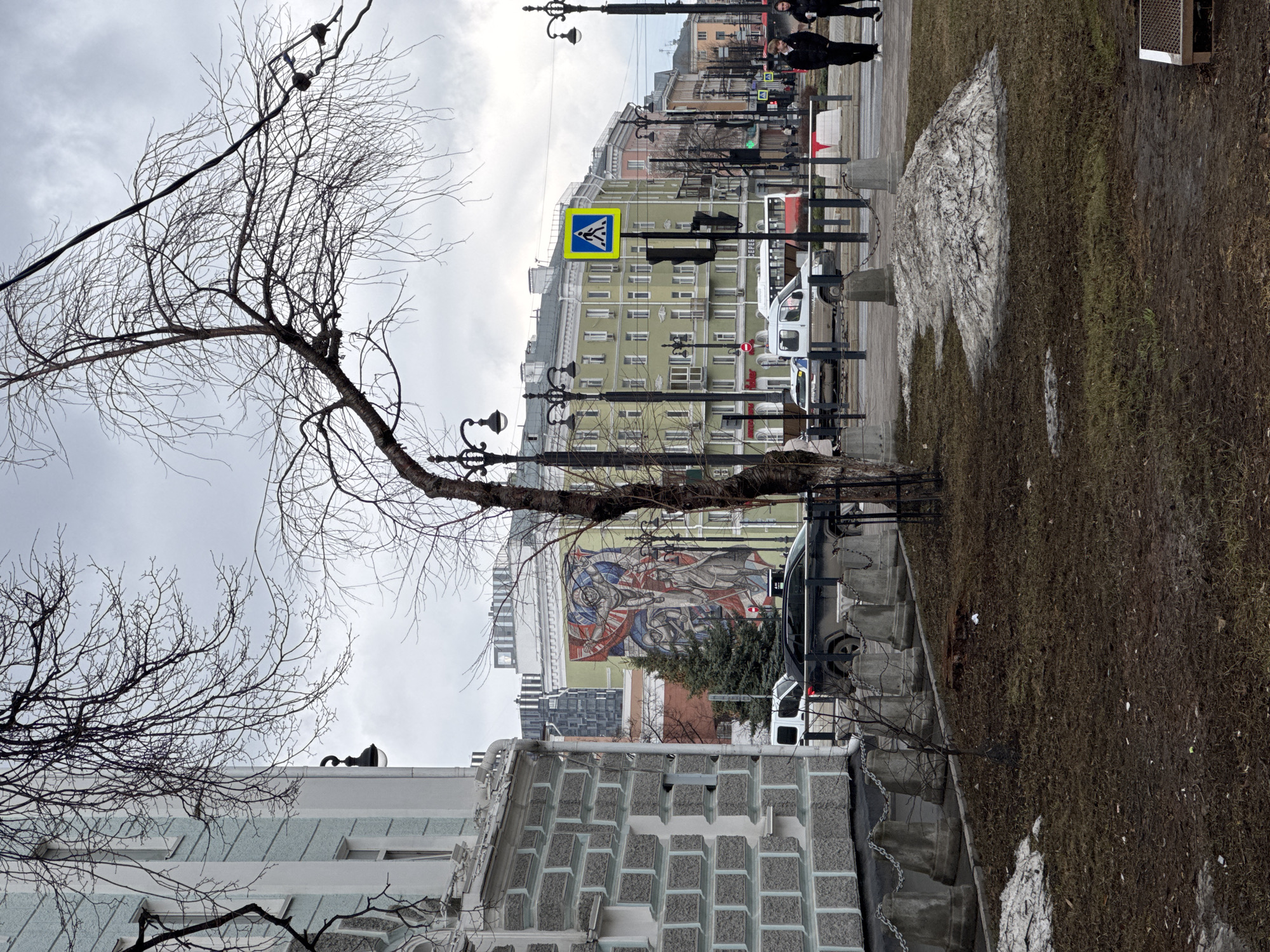 The image depicts a street scene in Perm, Russia, characterized by an overcast sky. The foreground includes a leafless tree obscuring parts of the scene, along with a building with a stone-textured facade. A sidewalk runs along the right side of the image, with patches of melting snow and brown grass. A line of street lamps stands along the sidewalk. Vehicles, including vans, are parked along the street. In the background, buildings of varying heights line the street, some of which feature murals. Pedestrians are visible walking along the sidewalk in the distance. A pedestrian crossing sign is prominent among the street furniture.