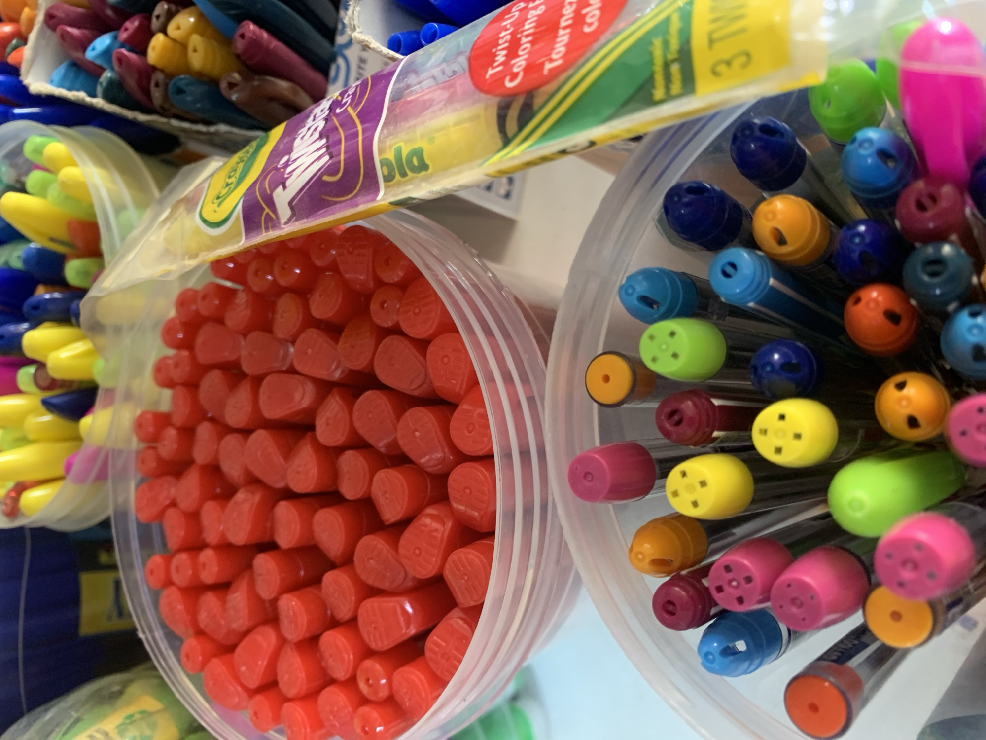 The image shows a close-up of several containers filled with colorful art supplies. In the foreground, a clear plastic container holds an assortment of markers with brightly colored caps, including pink, yellow, green, orange, and blue. Behind this, another clear plastic container is filled with red cylindrical objects, possibly crayons or markers. To the right, a partially visible package displays the "Crayola Twisty" logo and text indicating "Twist-Up Coloring Eggs" and "Tournez..." The background reveals more art supplies, including yellow and pink objects in a clear container, and a darker container with a blue lid. The overall scene suggests a retail environment or a well-stocked art supply collection. The lighting appears to be overhead and bright, casting subtle shadows. There are no people visible in the image.