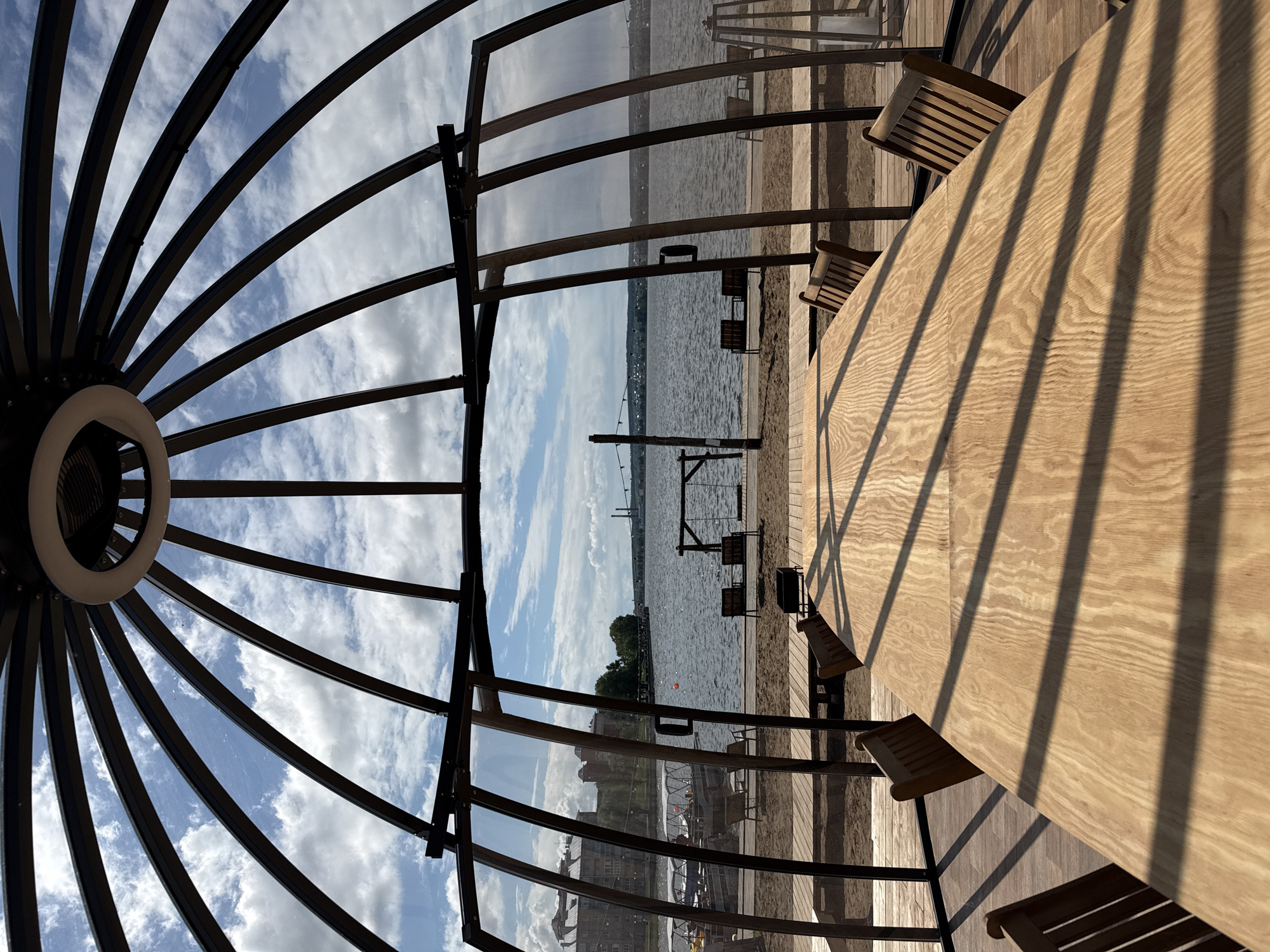 The image shows an outdoor scene in Votkinsk, Russia, featuring an outdoor structure with a dome-like roof. The structure encloses a round wooden table with several chairs positioned around it. The structure has glass panels, offering a view of a beach area and a body of water, possibly a river or lake. In the distance, there is a building on the shoreline and some vegetation. There are also structures and furniture on the beach. The photograph appears to have been taken during the day.