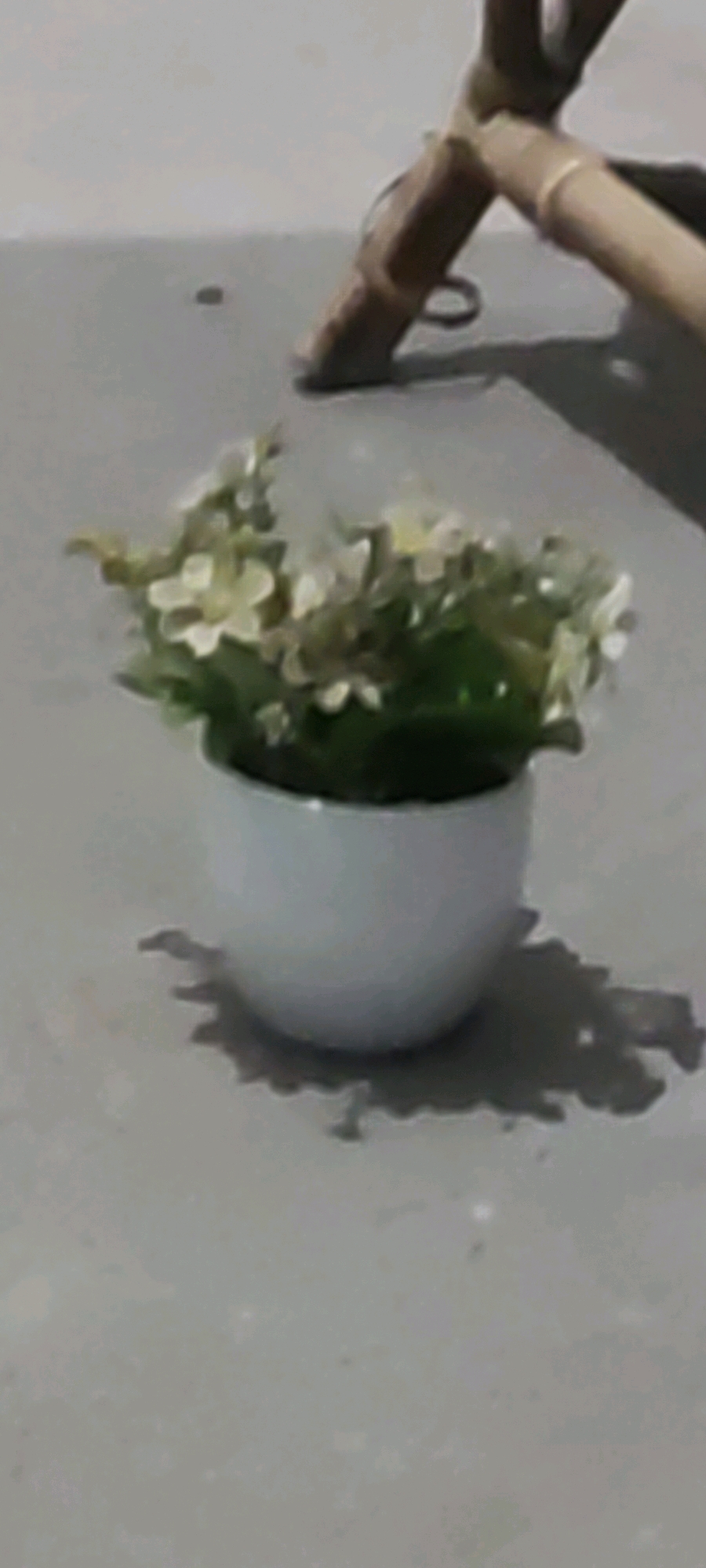In Karangwangun, Indonesia, an artificial plant with small white flowers sits in a white pot on a gray, textured surface. A portion of a wooden structure, possibly part of a chair or table leg, is visible in the upper right portion of the frame, casting a shadow on the surface. The lighting suggests it is daytime.