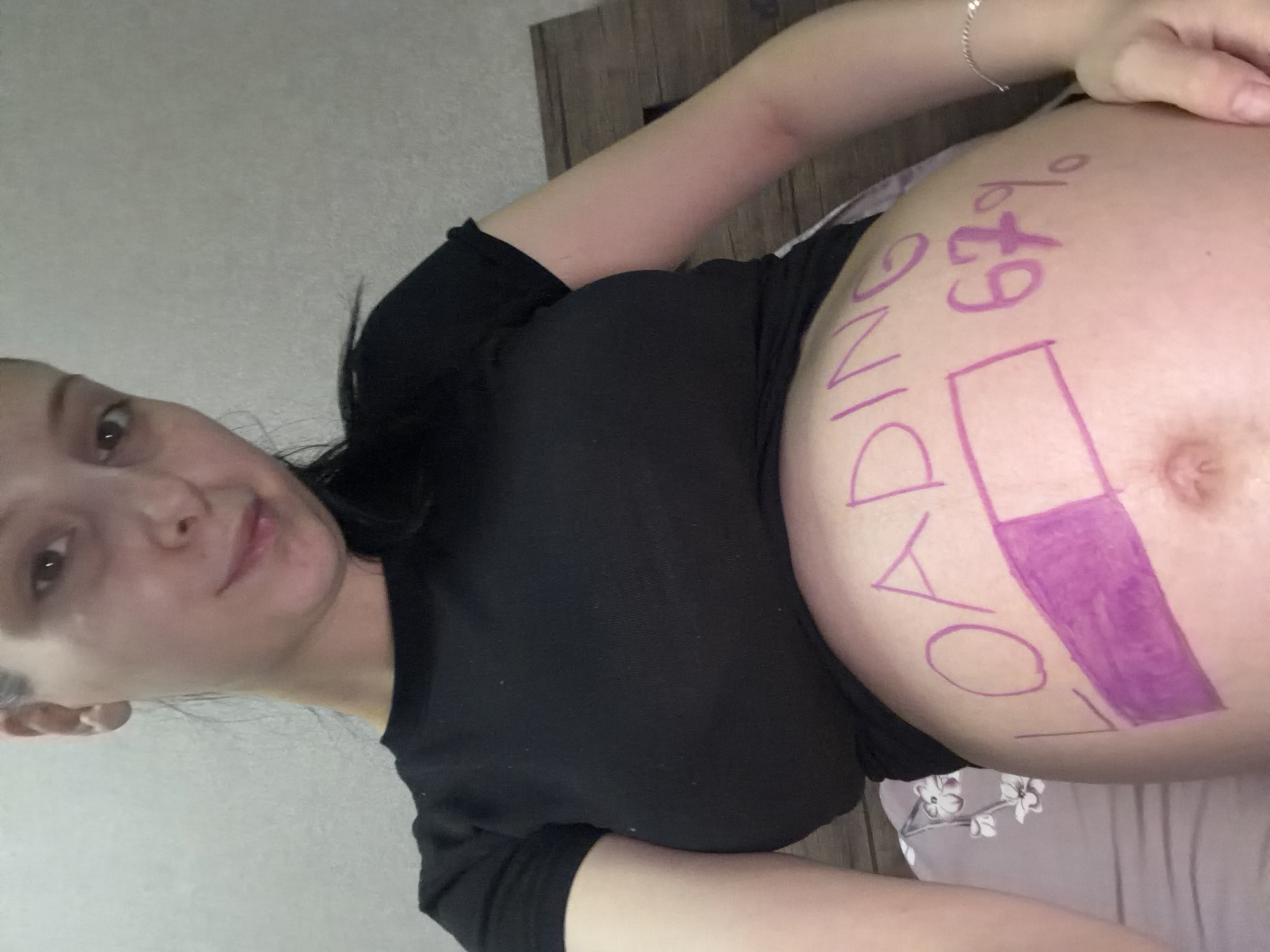 A woman, likely pregnant, is pictured in a close-up shot. She is wearing a black top and has writing and a drawing on her belly. The text reads "LOADING 67%" and there is a purple rectangle next to it, resembling a loading bar, with the purple filling about two-thirds of the rectangle. The woman is looking towards the camera with a slight smile. The background appears to be a wall and a wooden surface, possibly furniture. The lighting suggests it could be daytime. The overall tone of the image is lighthearted and celebratory, likely indicating the progress of a pregnancy. There are no other significant objects or people visible in the frame. The location is specified as Tbilisi, Georgia, though this context is not visually apparent in the image itself.