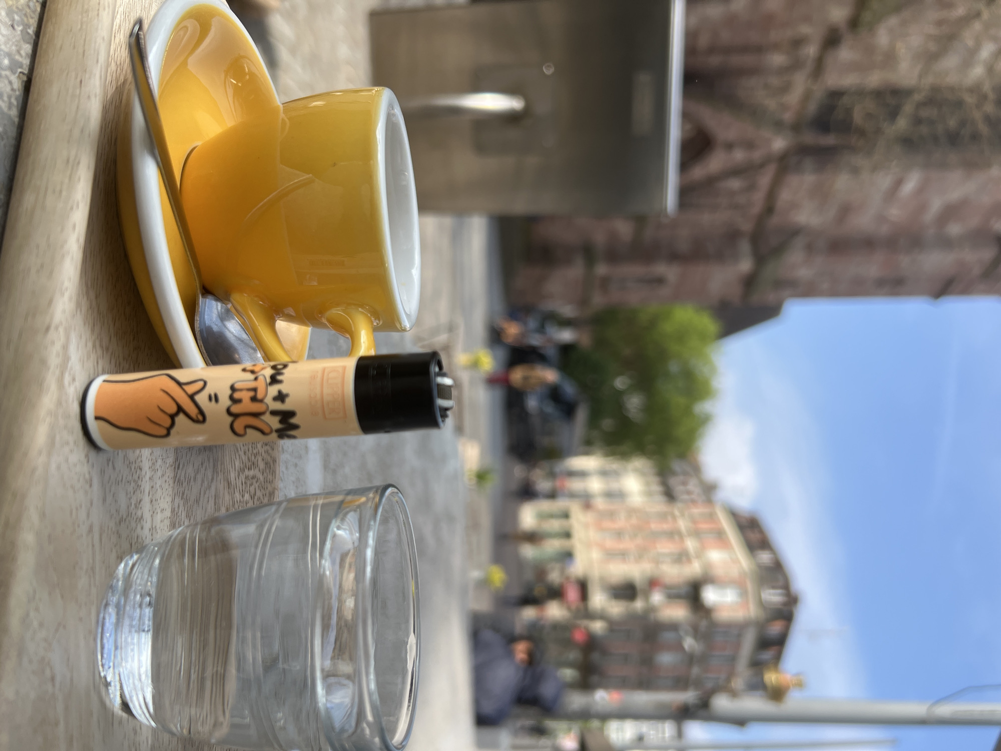 The photograph is taken at a table at an outdoor cafe in Strasbourg, France. A yellow teacup and saucer with a spoon are on the table. A clear glass of water and a lighter with a cartoon hand design are also visible. In the blurred background, people are walking on the sidewalk, and buildings are visible. The sky is blue.