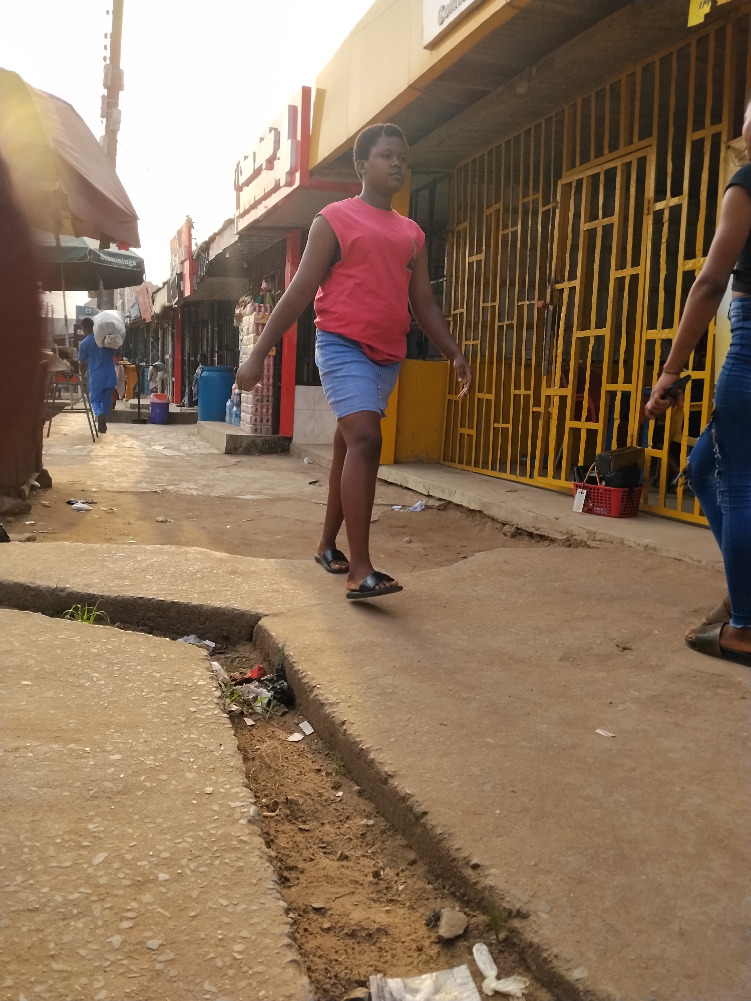 The image depicts a street scene in Ura Nta Umuarandu, Nigeria. A person in a pink sleeveless shirt and blue shorts walks on a paved area. To the right is another person in blue jeans. Buildings line the street with yellow metal gates. A red plastic basket sits next to the gates. Debris litters the edge of the pavement. In the distance, another person is carrying a white sack on their shoulder. A green umbrella with "Seasonings" written on it is also visible.