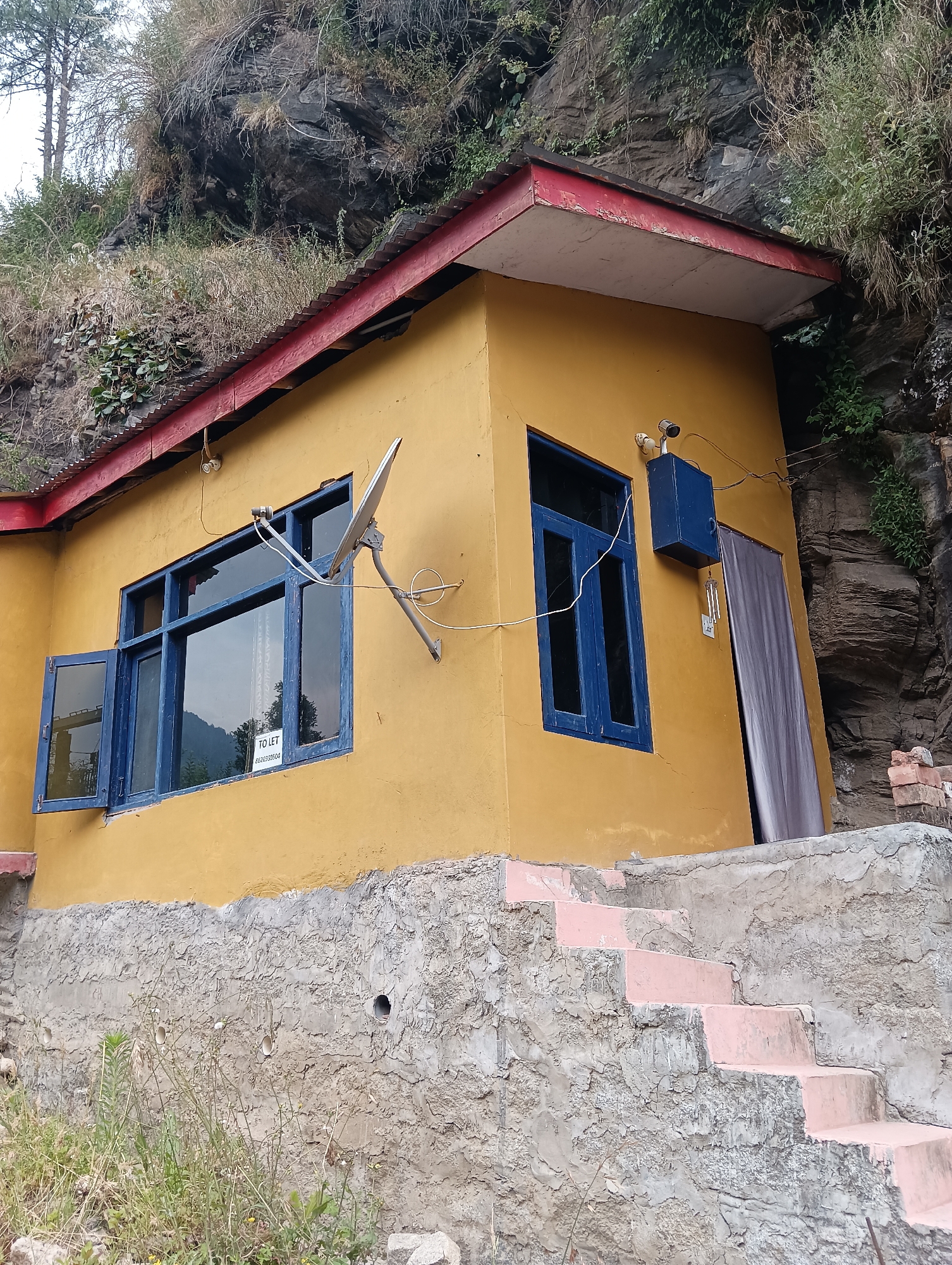 The image shows a yellow house built into a rocky hillside in Old Manali, India. The house has a red-trimmed roof and blue-framed windows. A satellite dish is mounted on the exterior wall next to a window. There is a blue electrical box above a doorway to the right of the window. A set of concrete stairs, with pink accents, leads up to the house from the foreground. The stairs and the lower portion of the house are surrounded by rough, textured concrete. Vegetation, including grass and some trees, is visible on the hillside and around the base of the house. The rocky hillside behind the house is covered in sparse vegetation. There are no people or animals visible in the image.